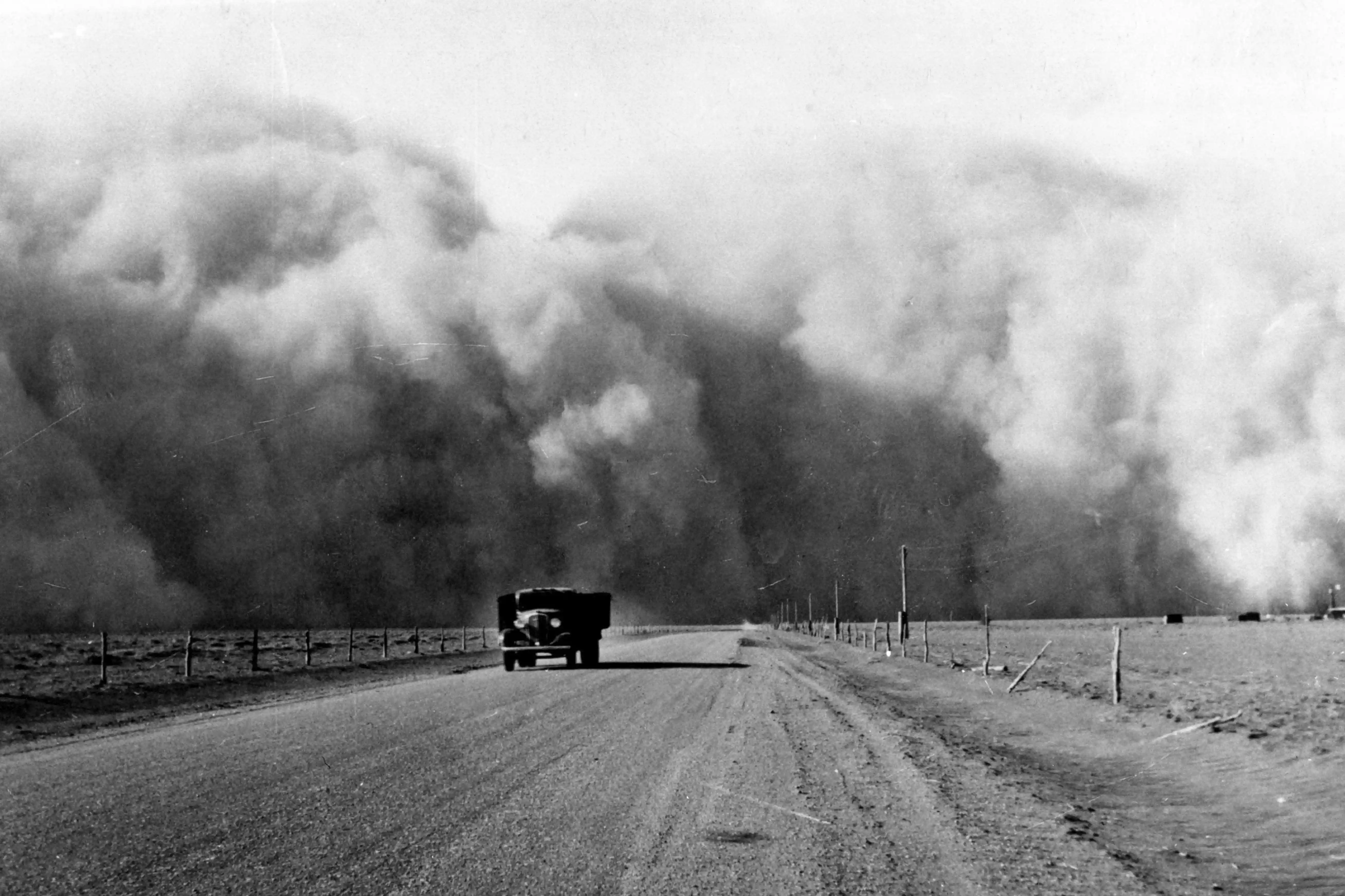 Dust cloud fills the sky and truck drives on dirt road away from it