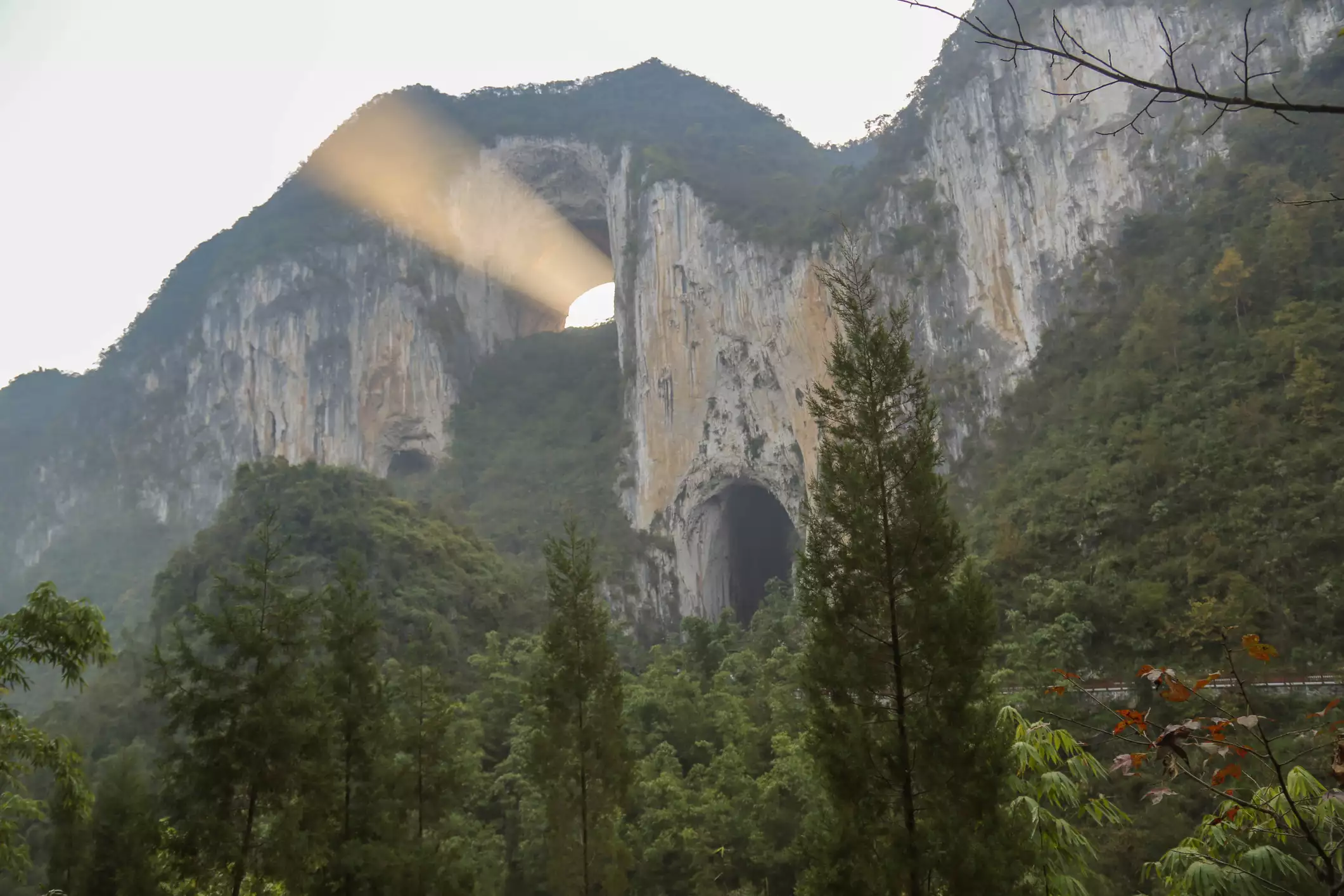 A forested mountain ridge with a natural arch and a beam of sunlight coming from the arch