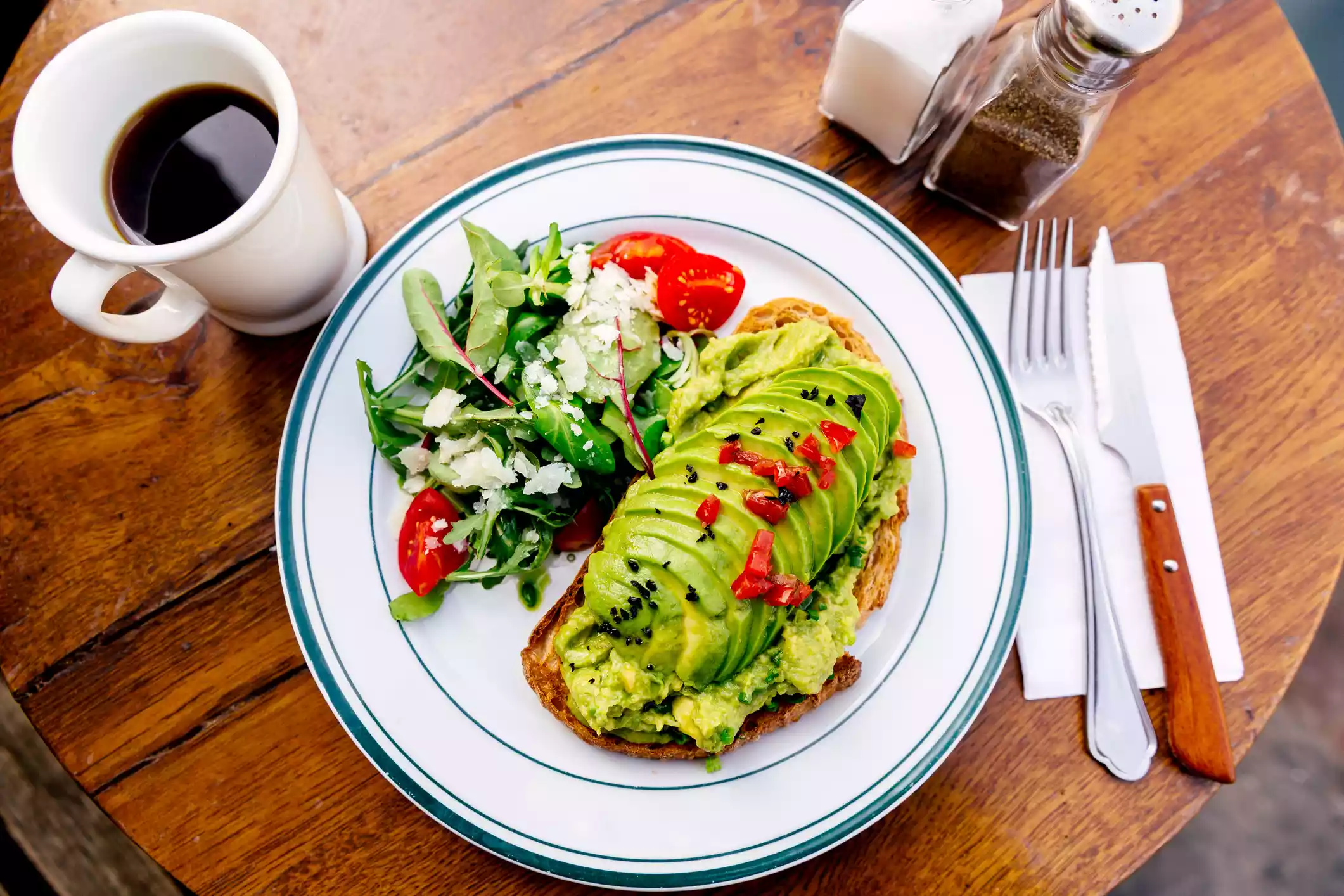 Overhead view of a small, round wood table with a white plate with blue trim filled with a large serving of avocado toast with a green side salad next to a mug of black coffee with salt and pepper shakers and a knife and fork on a napkin 