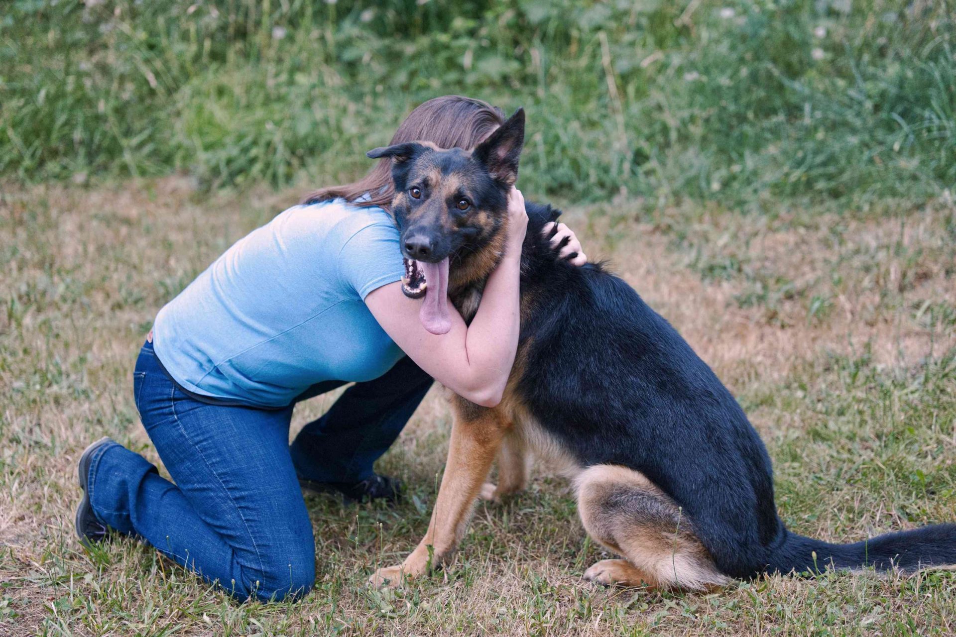 una mujer se agacha hasta el suelo para abrazar a un perro pastor alemán con la lengua fuera