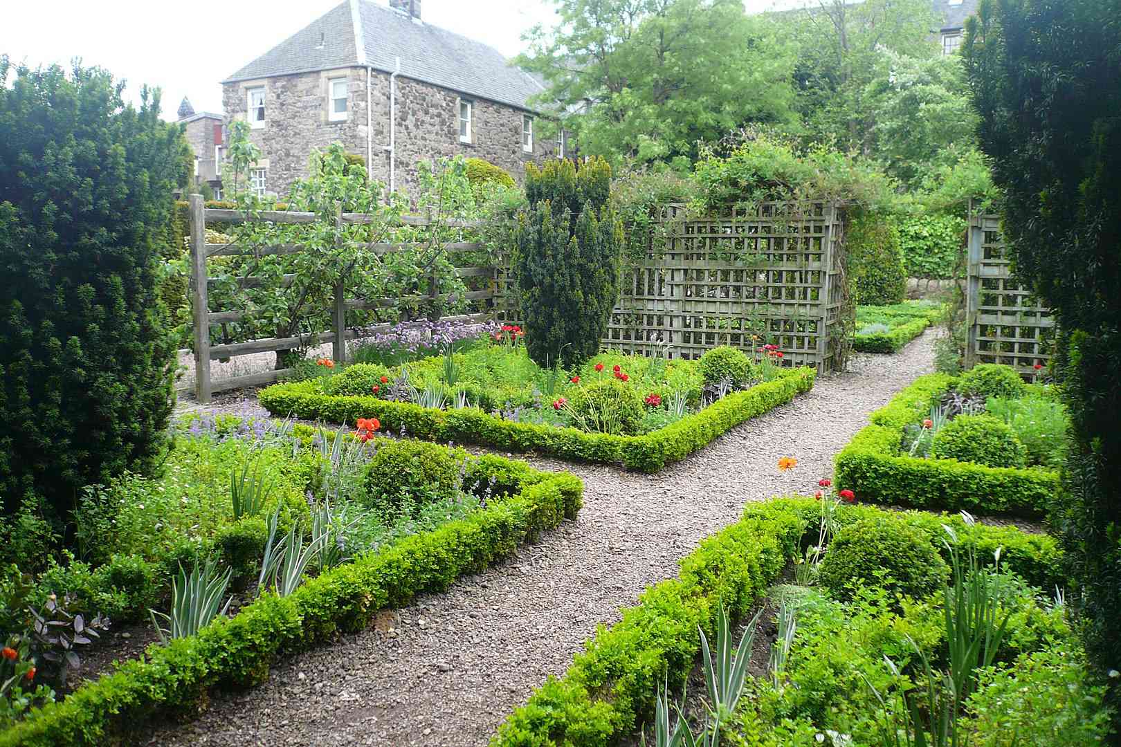 Caminos a través del jardín de Dunbar's Close, un jardín ordenado con pequeños parterres de plantas verdes y arbustos de formas altas, con un enrejado de madera y una pequeña iglesia en la distancia