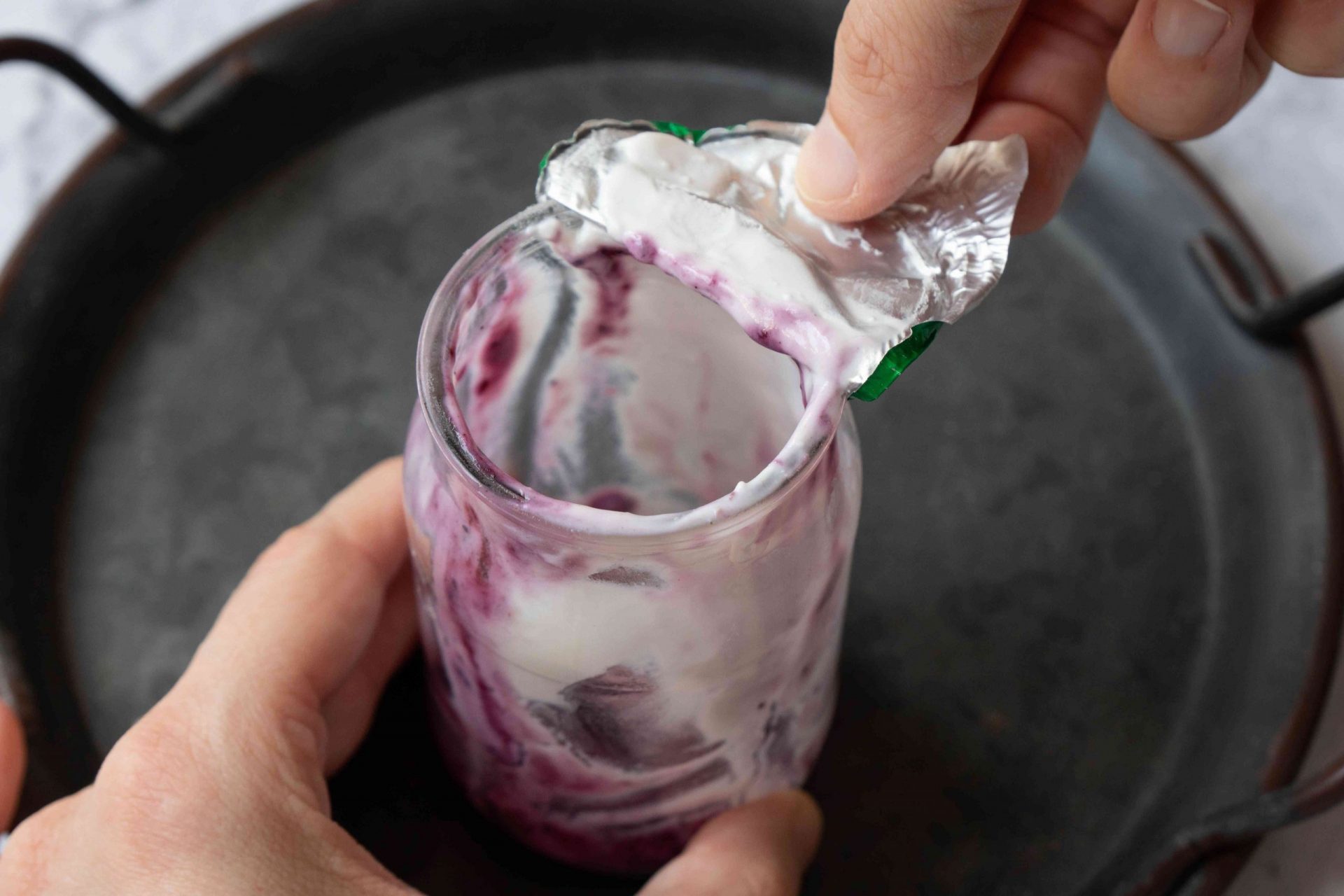 las manos quitan la tapa de papel de aluminio del envase de vidrio del yogur de fruta a medio comer