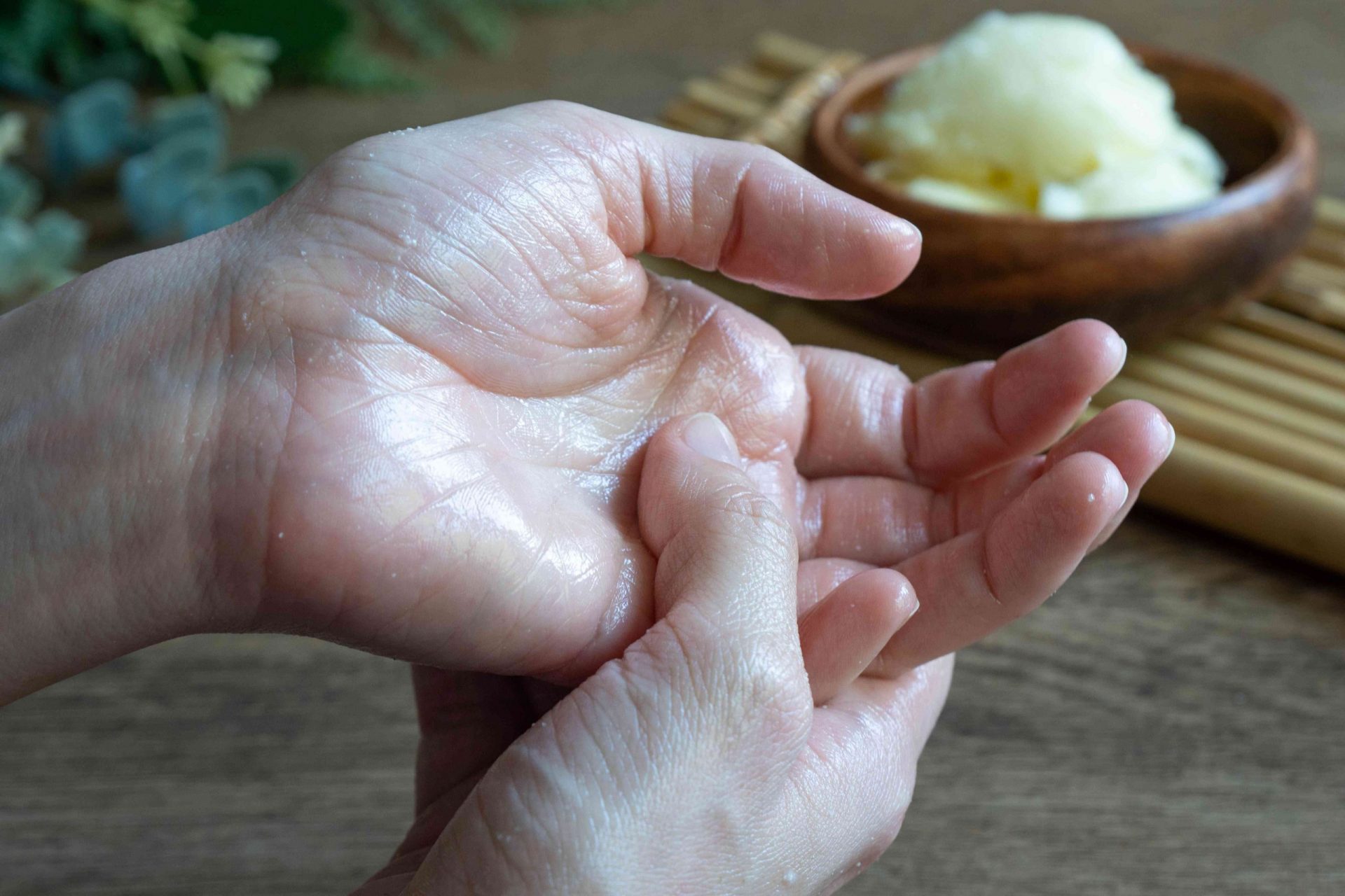 dos manos frotando manteca de karité en la palma de la mano, con un cuenco de madera con manteca de karité cruda de fondo