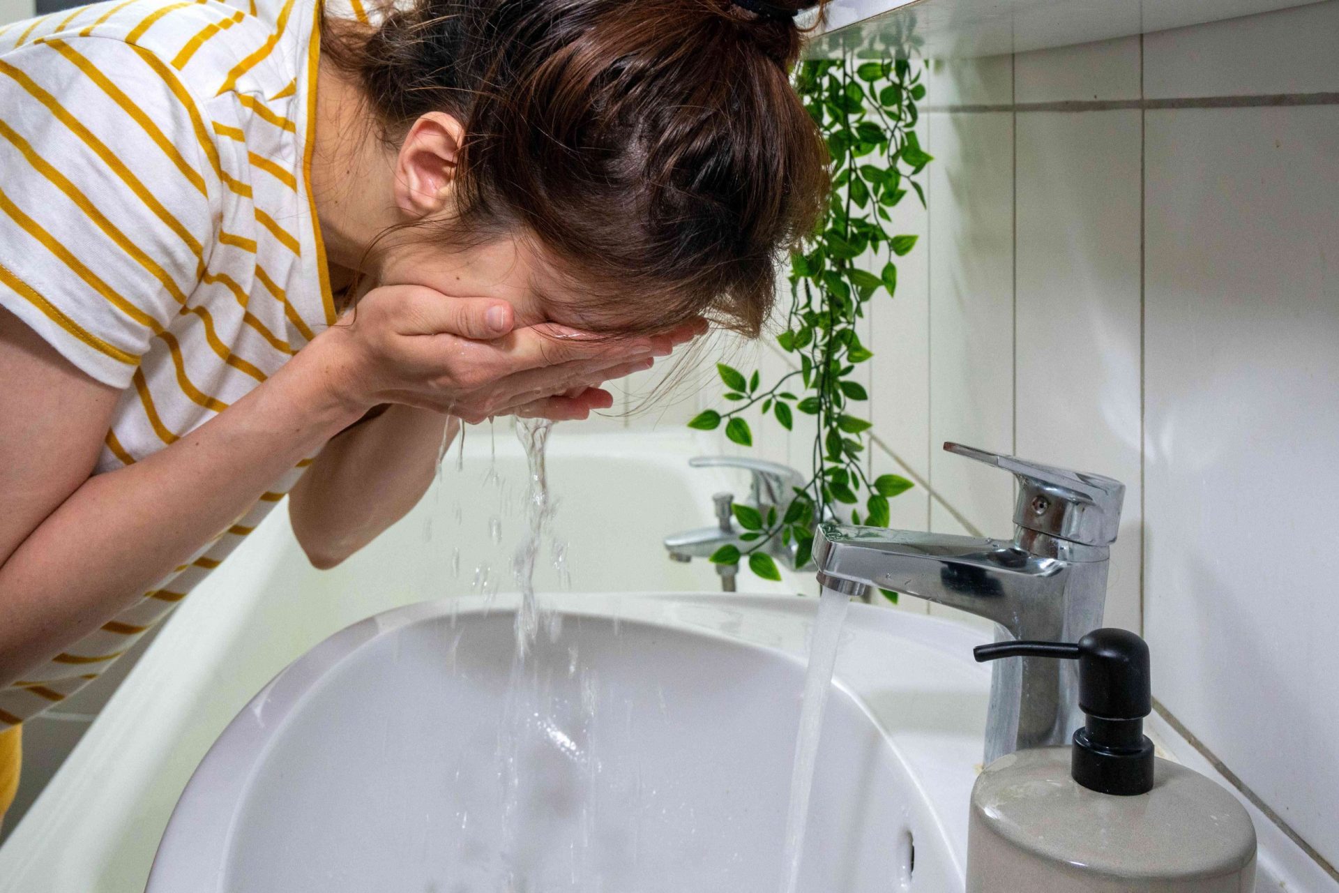 mujer con camisa de rayas se lava la cara en el lavabo del baño junto a una planta colgante
