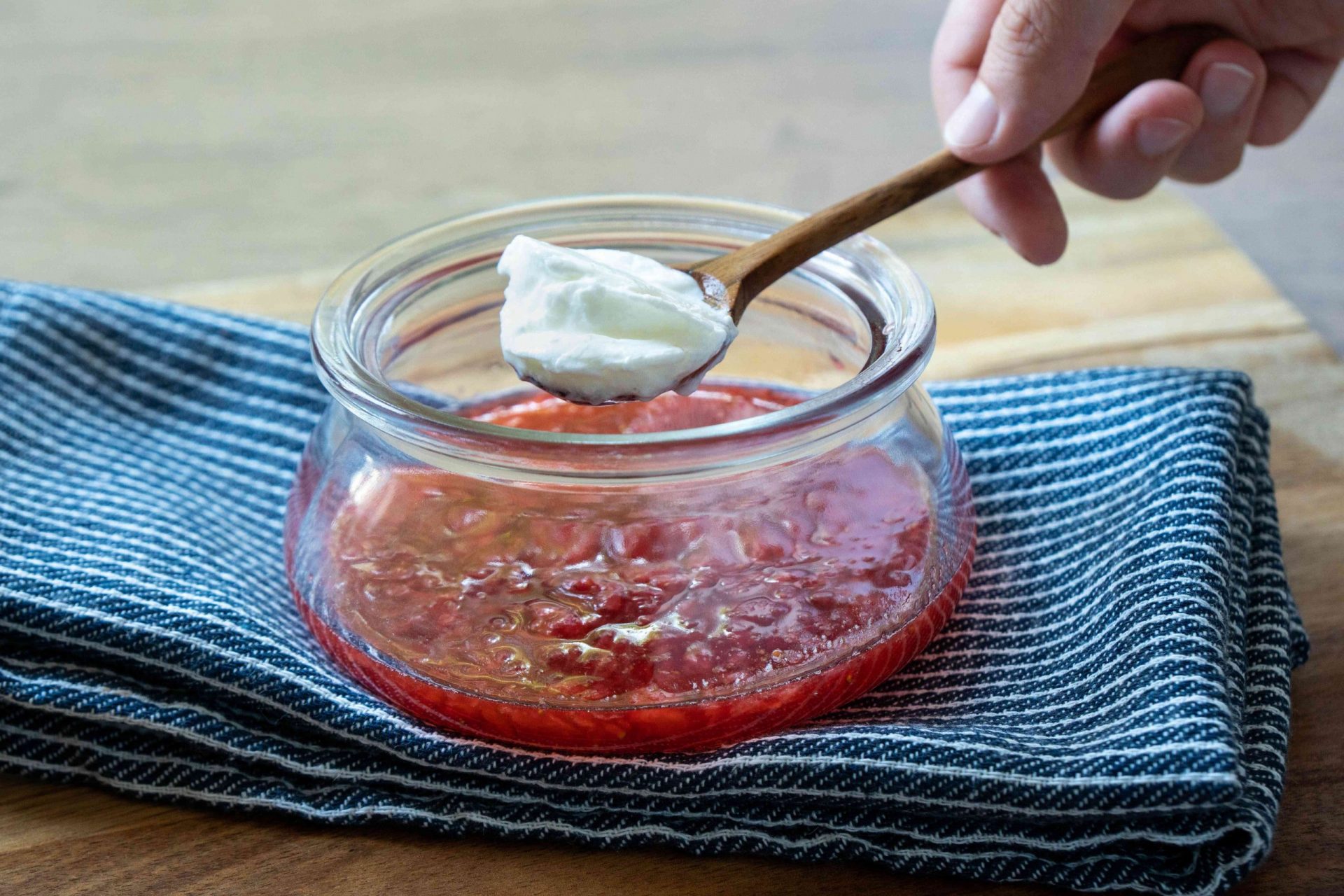 la mano añade una cucharada de yogur griego en una cuchara de madera a la mezcla de fresas trituradas en un bol de cristal
