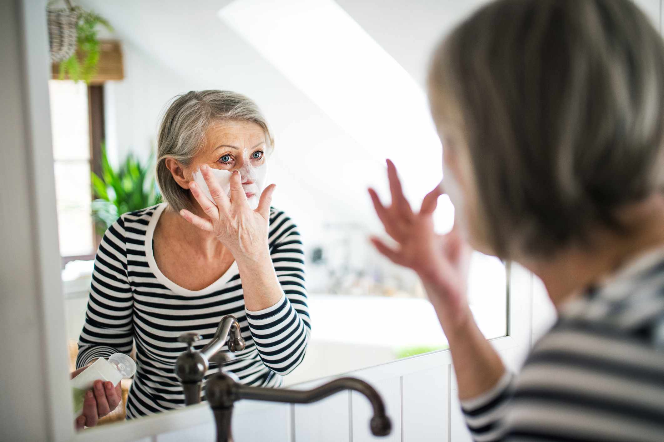 Una mujer mayor con camisa de rayas se aplica una mascarilla facial en el espejo