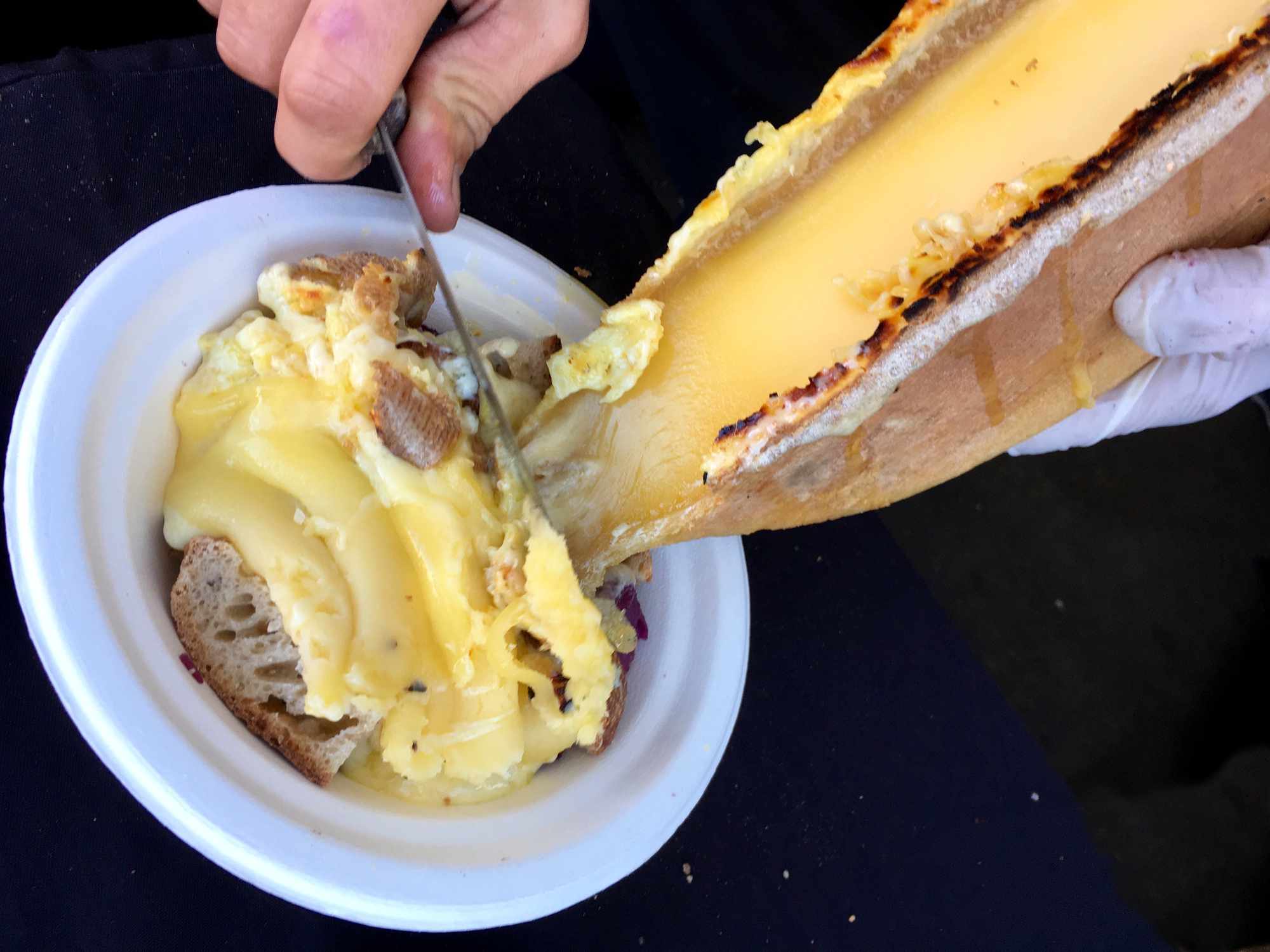 Queso raclette derretido siendo raspado de una rueda de queso sobre un bol de cubos de pan y patatas