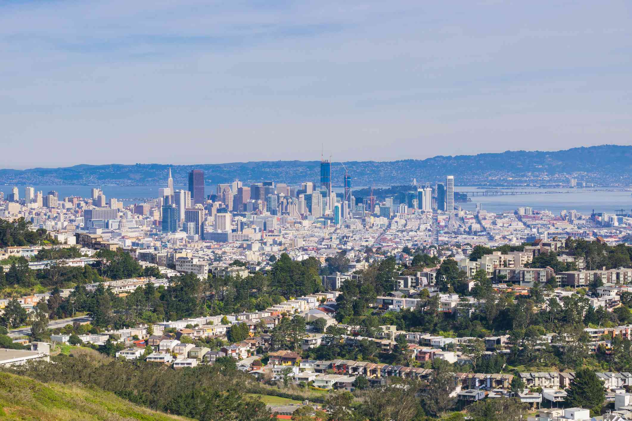 Vista de la ciudad de San Francisco desde el monte Davidson, con el cielo azul y montañas más pequeñas en la distancia, los altos edificios del centro de la ciudad y edificios más pequeños en primer plano situados entre altos árboles verdes