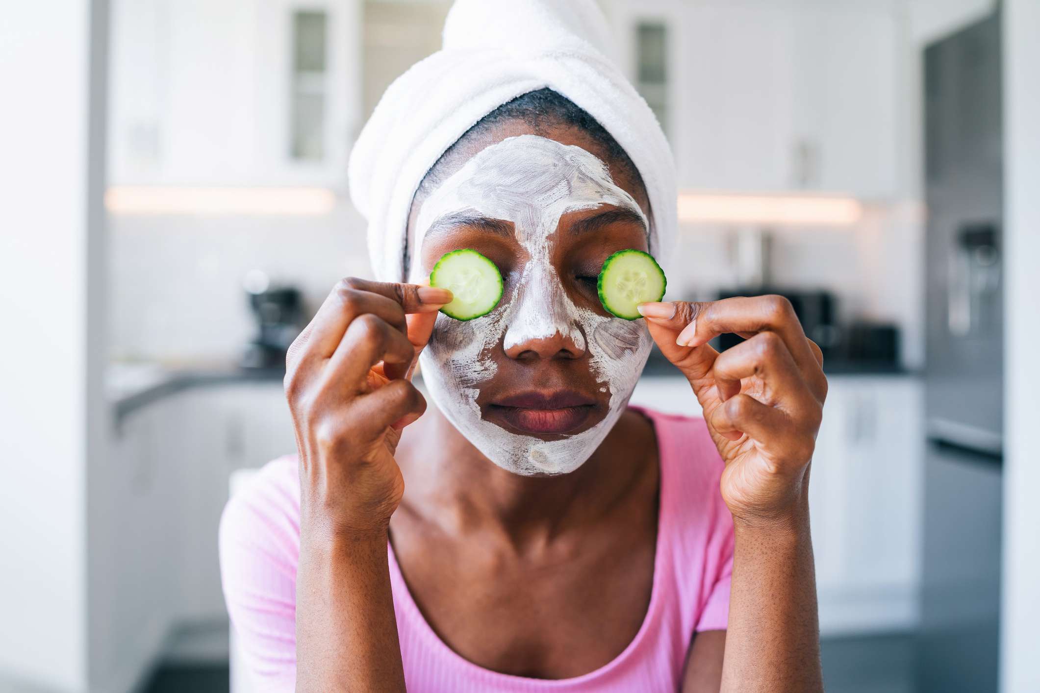 Una mujer negra con una máscara facial y sujetando pepinos en los ojos