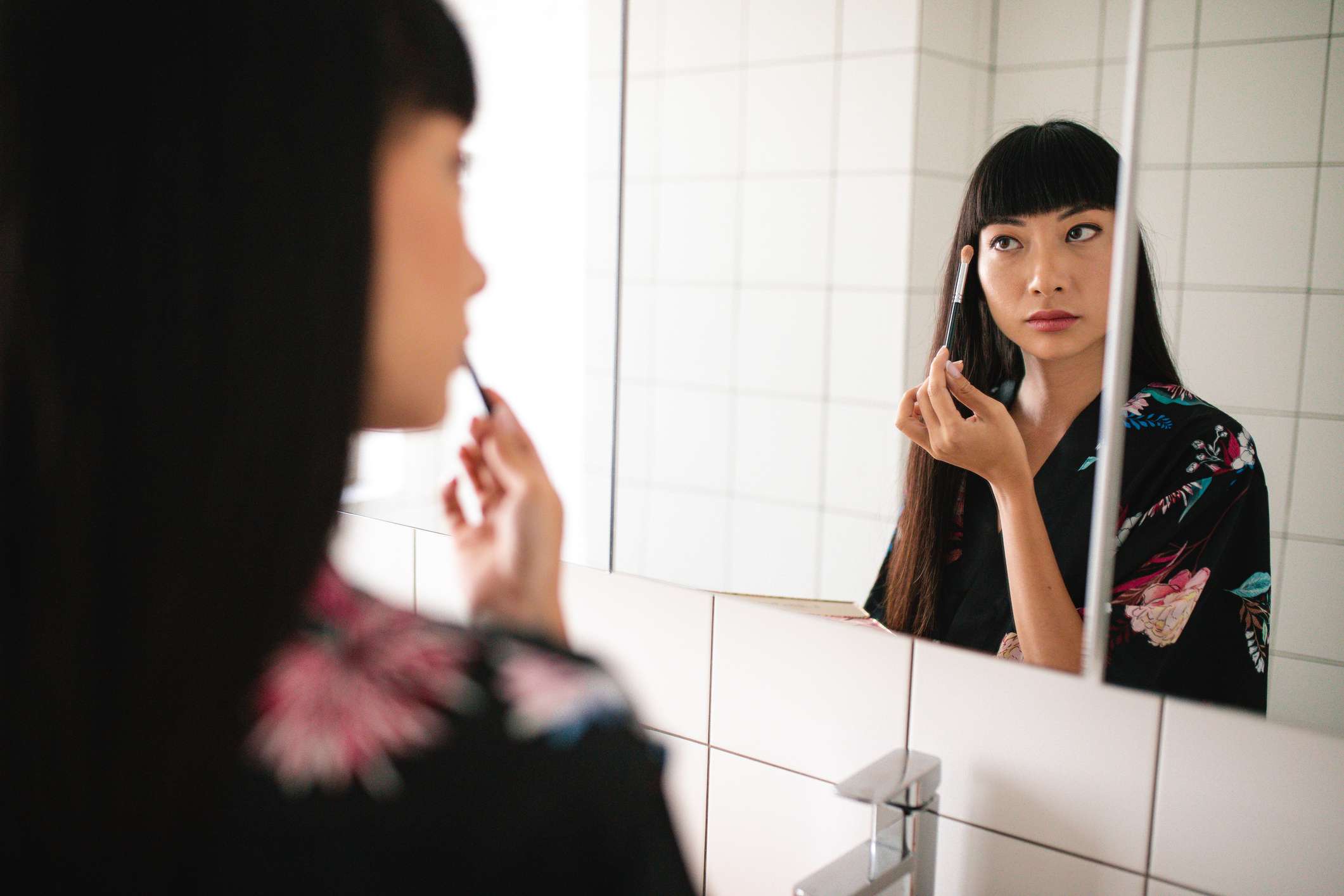 Una mujer asiática con flequillo y pelo largo se aplica la sombra de ojos en el espejo