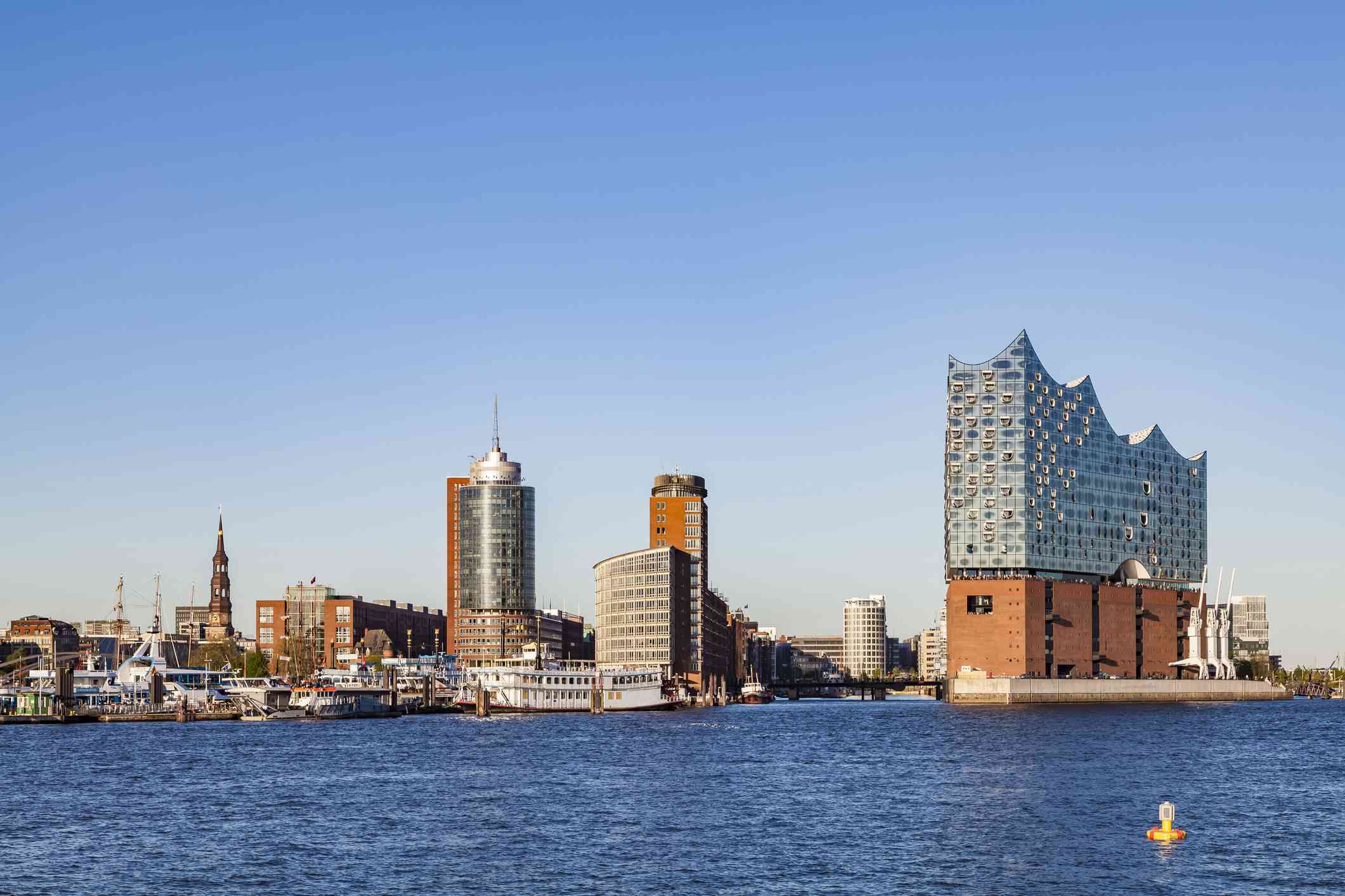 vista a través del agua azul del puerto de la elbphilharmonie de Hamburgo, a la derecha, un edificio de usos múltiples terminado en 2017, con varias plantas de ladrillo en el nivel inferior y una moderna torre de cristal en la parte superior, junto a edificios nuevos y antiguos en el puerto de Hamburgo,