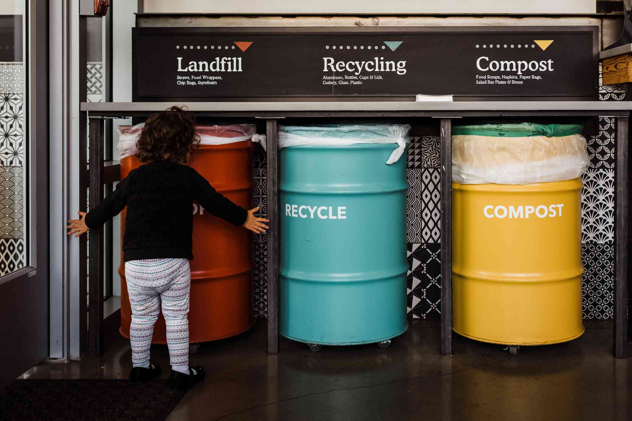 Niño pequeño delante de tres grandes contenedores etiquetados: vertedero (rojo), reciclaje (azul) y abono (amarillo)