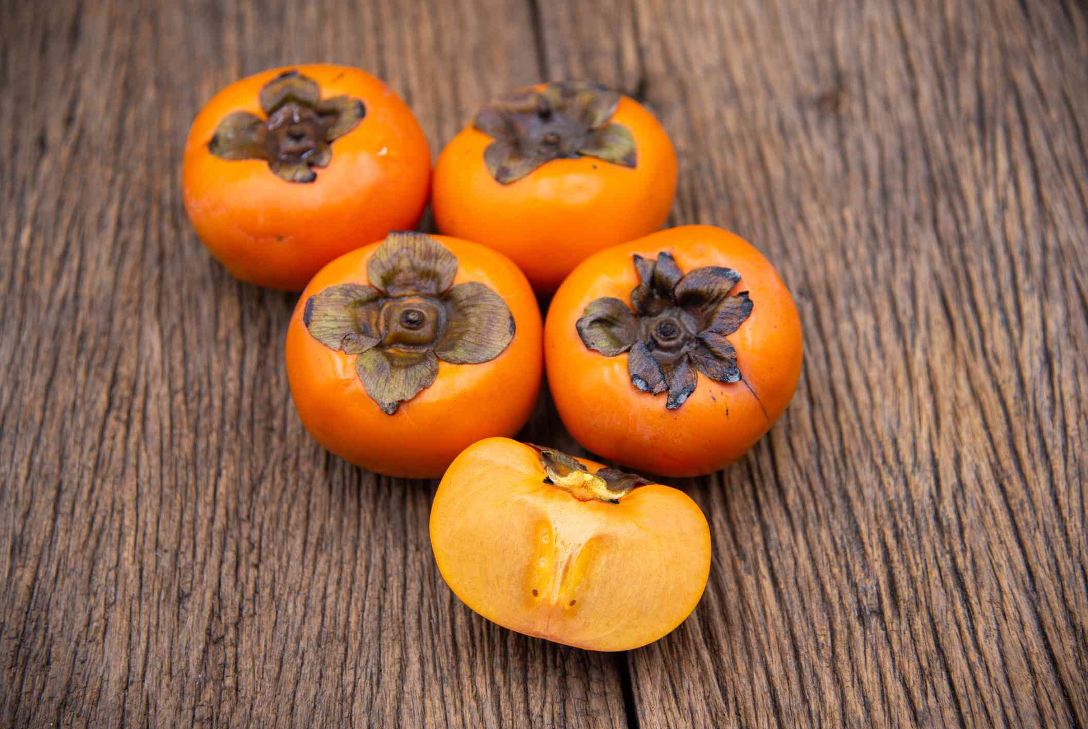 cinco caquis de color naranja Fuyu, uno de ellos cortado por la mitad, sobre una vieja mesa de madera