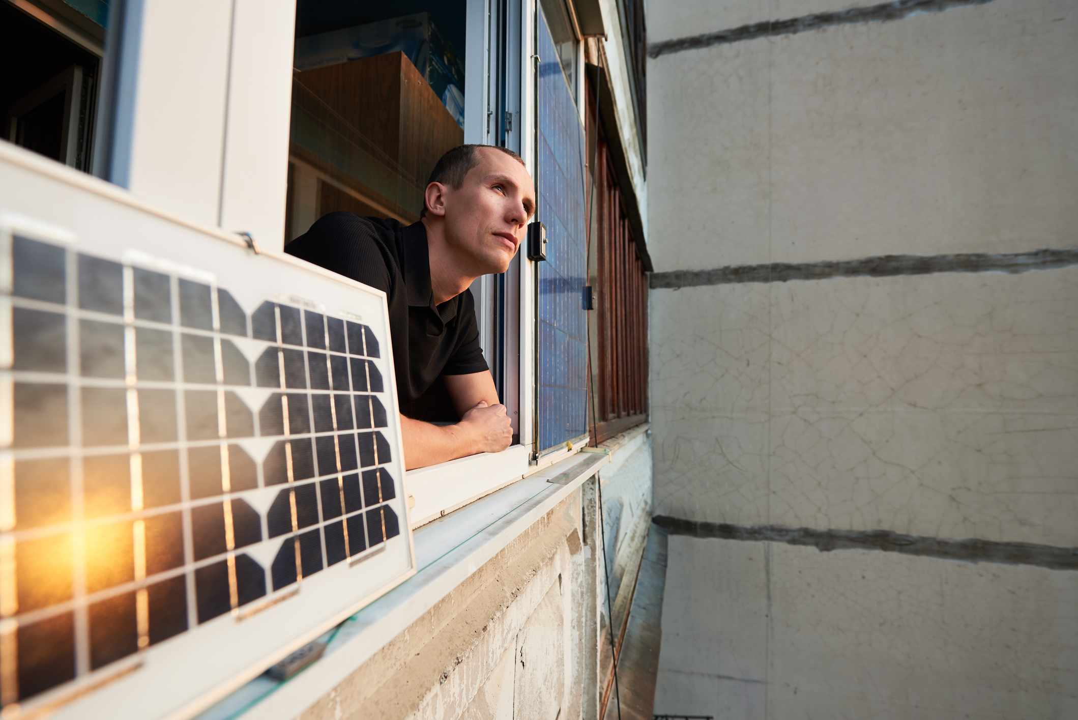 Un hombre se asoma a la ventana de un apartamento junto a un panel solar portátil fijado a la repisa de la ventana; el reflejo del sol es visible en el panel...