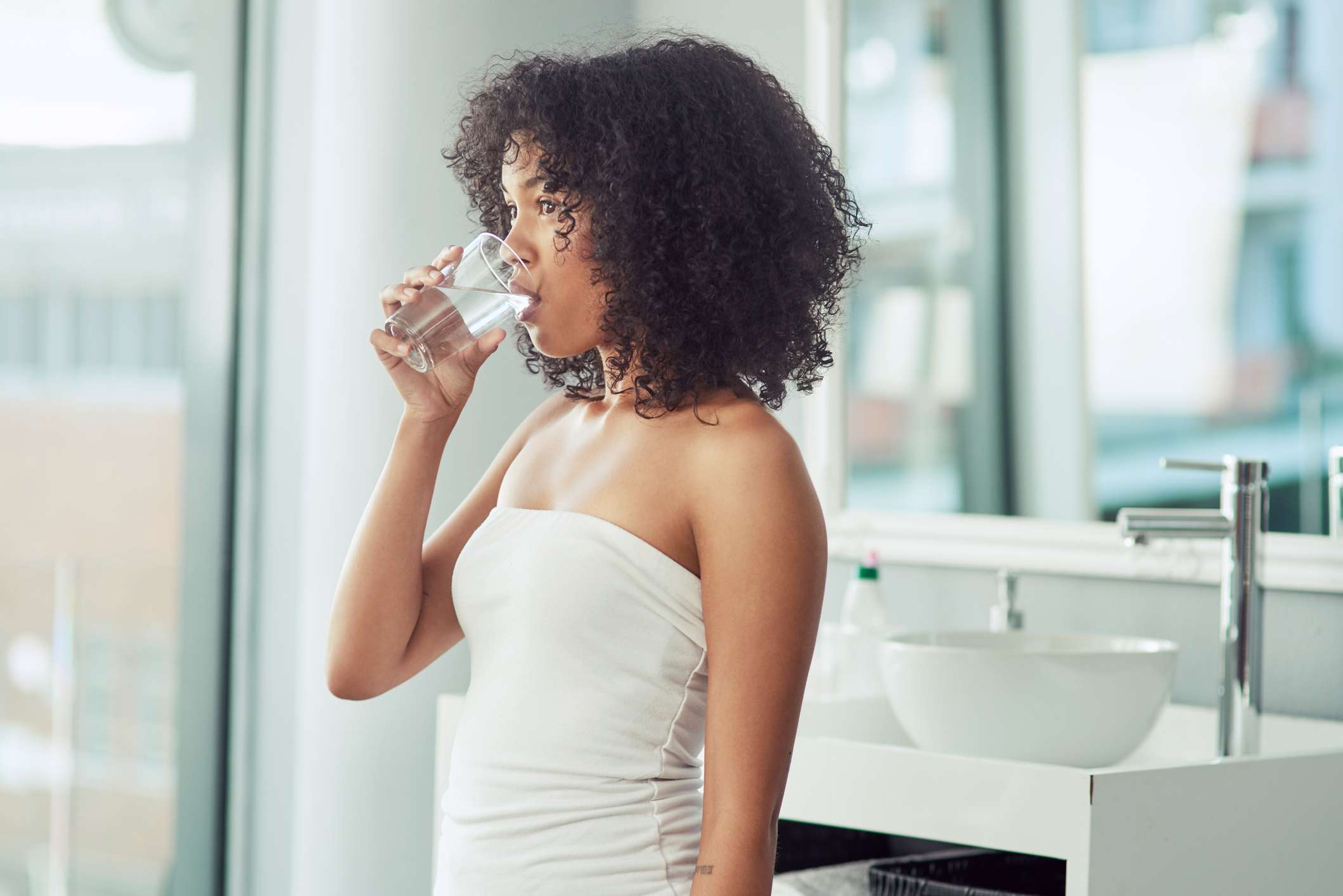Una joven negra bebe agua de un vaso en un lavabo blanco