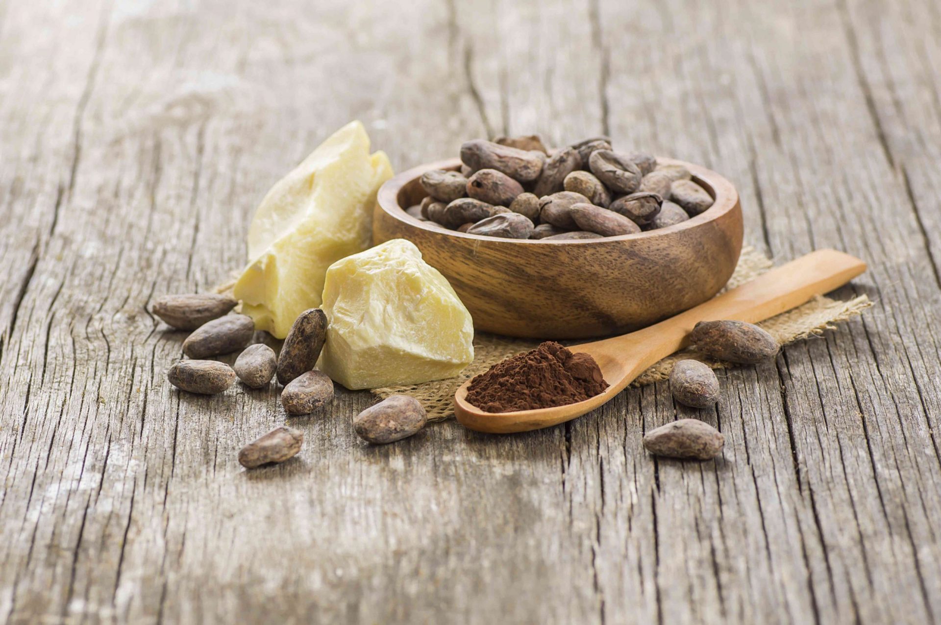 Manteca de cacao o aceite sólido de cacao con cacao en polvo en una cuchara y granos de cacao crudos en un cuenco de madera sobre un fondo rústico.
