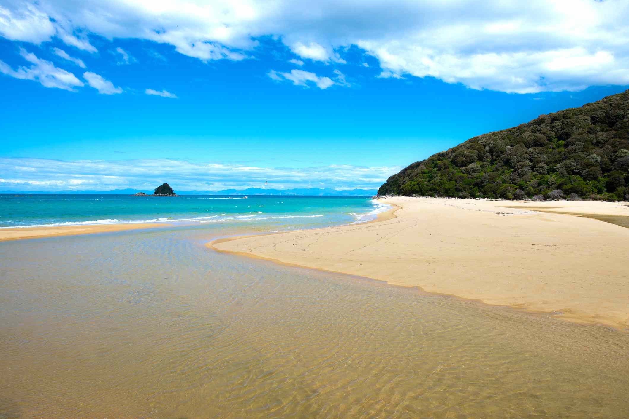 una amplia franja de playa de color crema a lo largo de un mar azul/verde con una exuberante montaña verde en la distancia y un cielo azul brillante con nubes altas en el Parque Nacional Abel Tasman