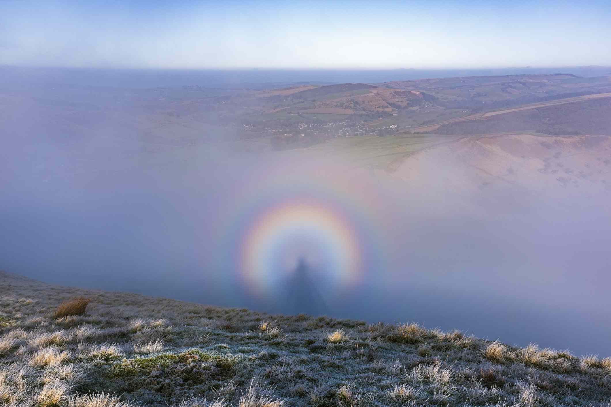 Una sombra ampliada de un observador proyectada sobre las nubes frente al sol, conocida como espectro de Brocken, parece una persona rodeada de un arco iris con montañas y un cielo azul/blanco encima