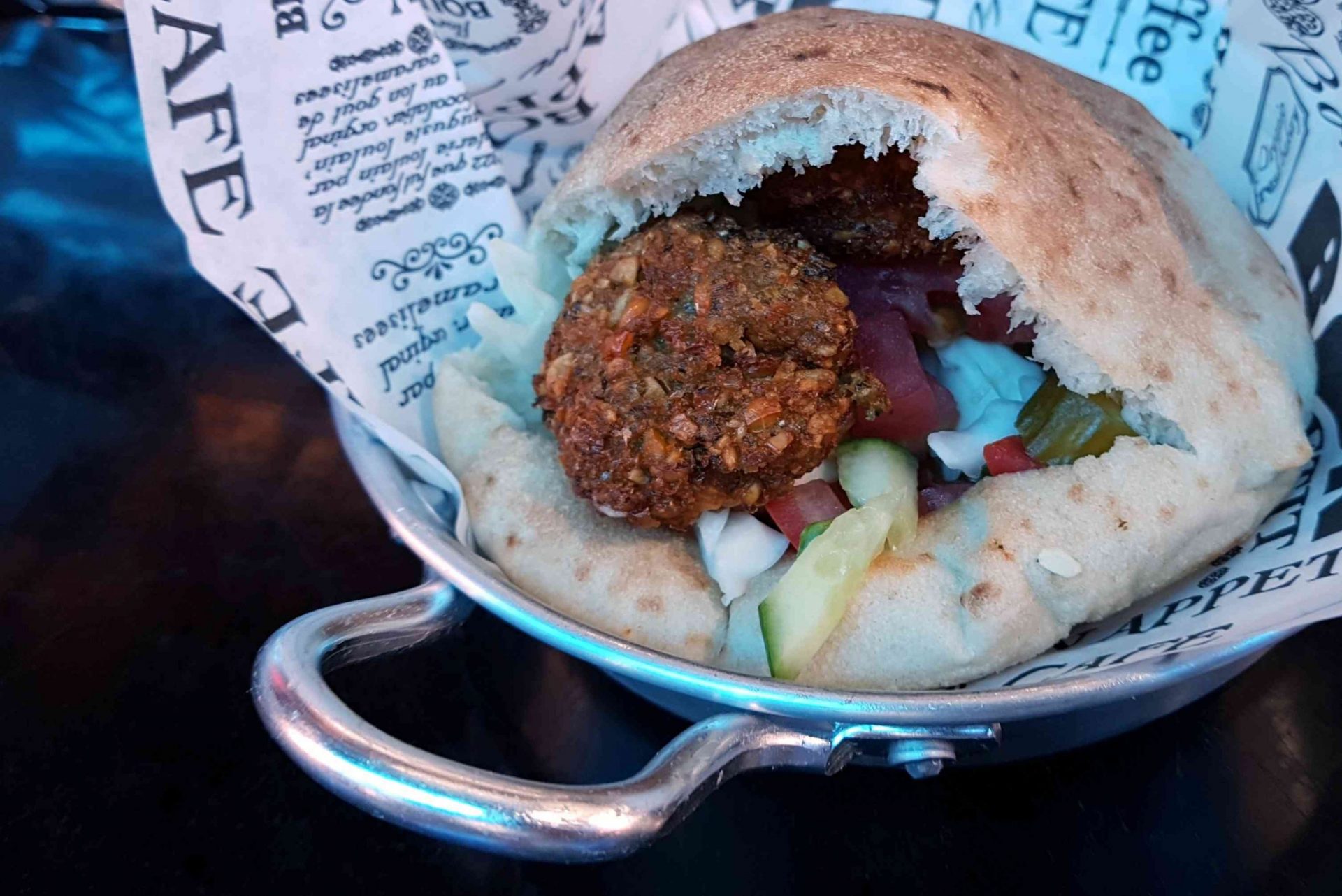 Un falafel kosher en una pita de bolsillo con rodajas de pepino y tomate servido en un cuenco de plata forrado con un menú de papel