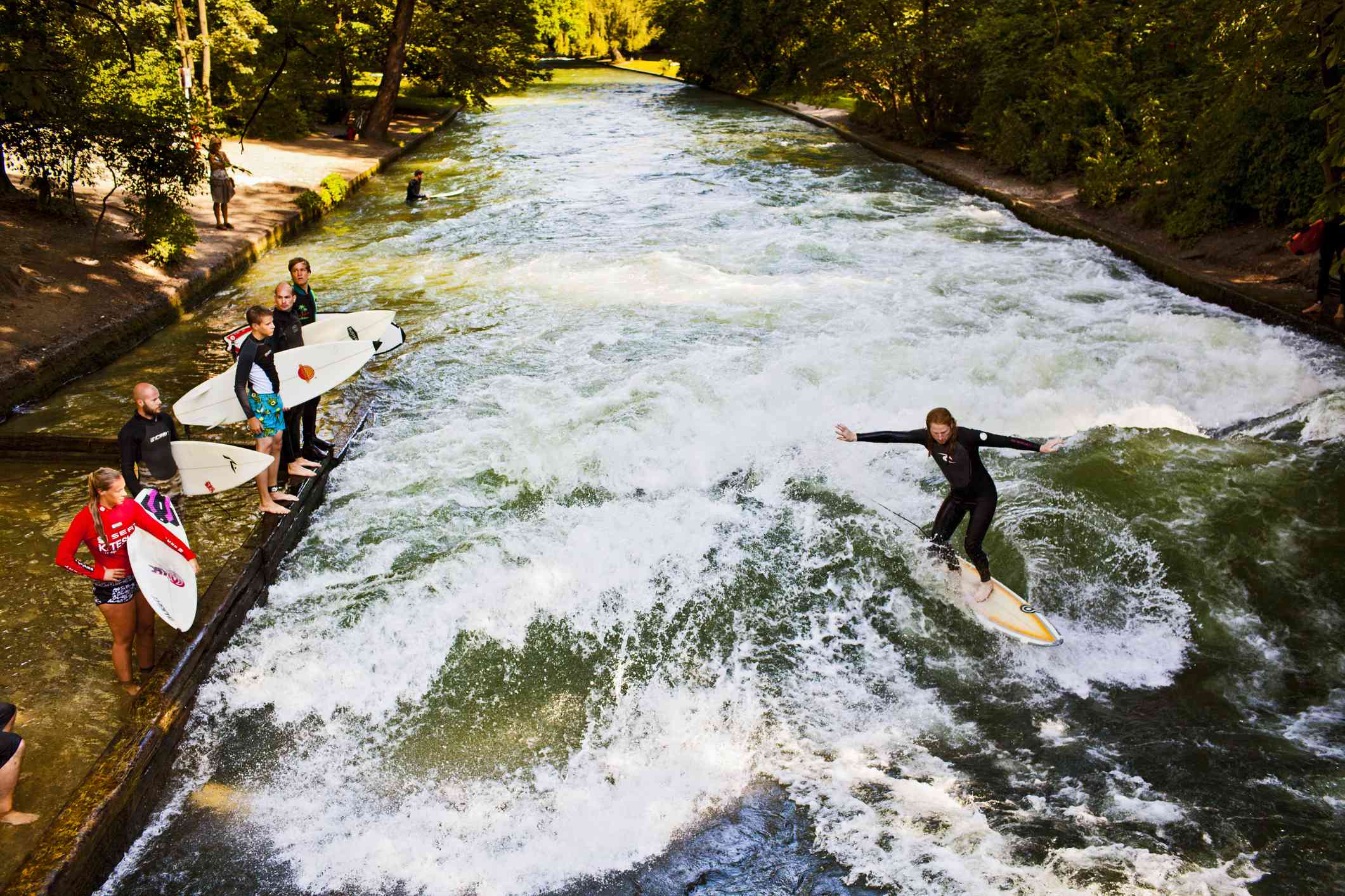 El agua verde y brillante del río Eisbach con un surfista montado en una moto mientras otros surfistas con sus tablas se sitúan en una plataforma cercana, más abajo del río, las orillas cubiertas de verde follaje en un día soleado