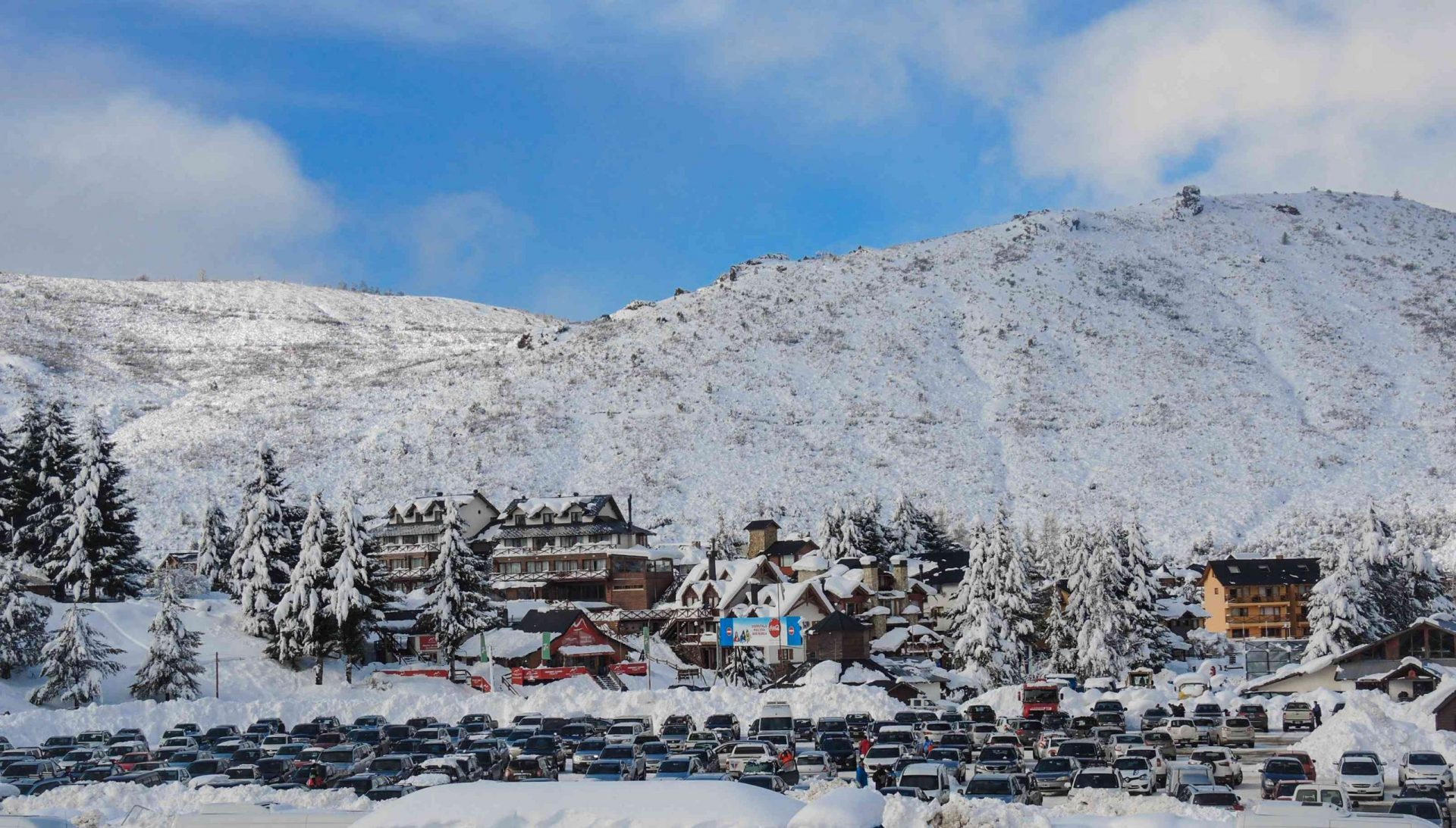La estación de esquí de San Carlos de Bariloche después de una nevada, el pintoresco pueblo, los árboles de hoja perenne y la montaña en la distancia están cubiertos de nieve bajo un cielo azul lleno de nubes blancas