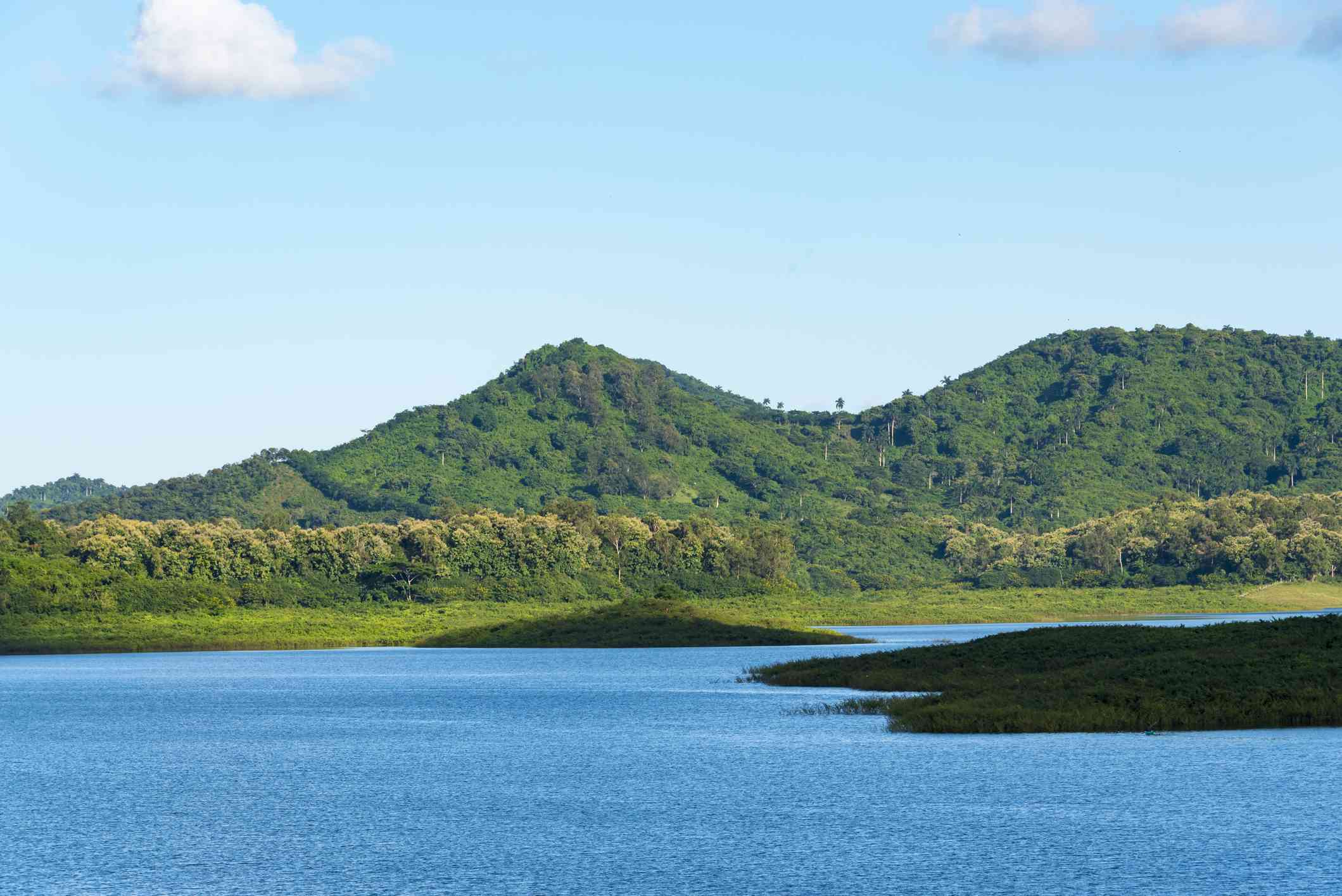 color azul vibrante del Lago de Hanabanilla rodeado de follaje verde bajo con las exuberantes montañas del Escambray cubiertas de verde en la distancia bajo un cielo azul con una ligera capa de nubes blancas