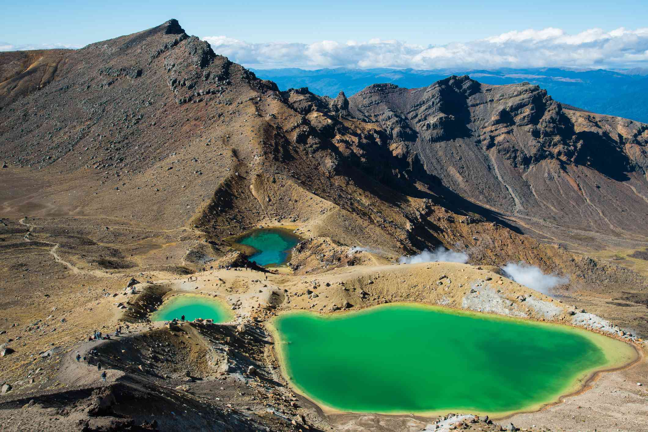 un lago grande y dos más pequeños de color verde esmeralda rodeados de montañas con un mar azul brillante de agua, cielo azul y nubes bajas y blancas en la distancia en el Cruce Alpino de Tongariro