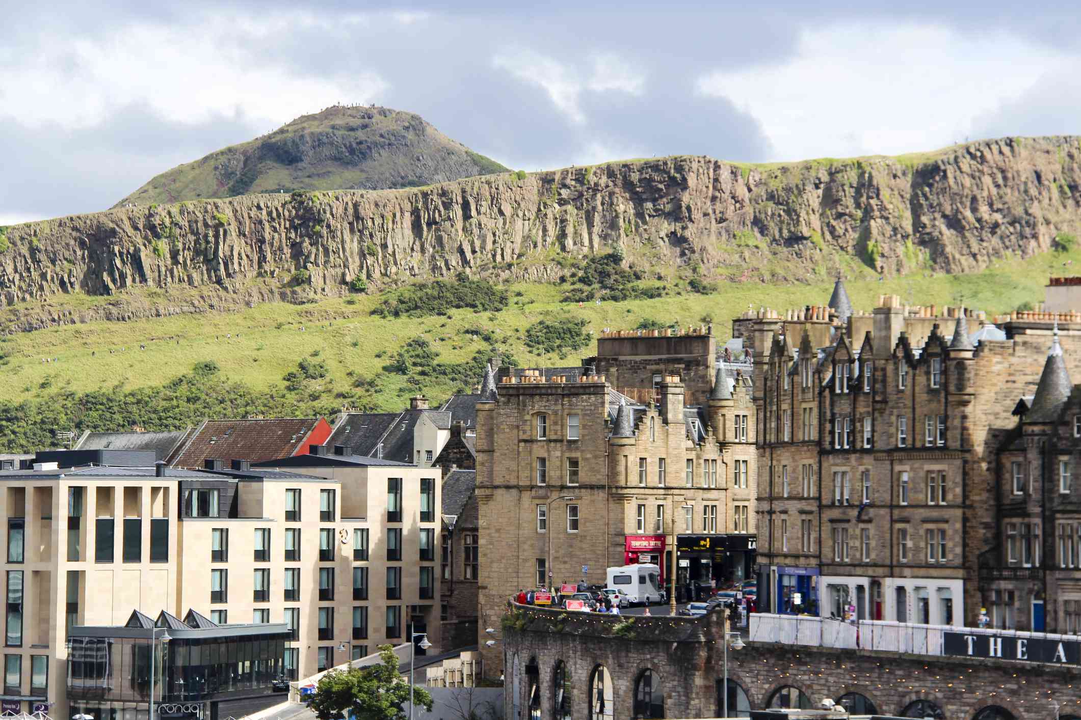 Vista de Arthur's Seat, una montaña de la ciudad de Edimburgo, Escocia, detrás de colinas más pequeñas y un campo cubierto de hierba con edificios de varios pisos de la ciudad en primer plano