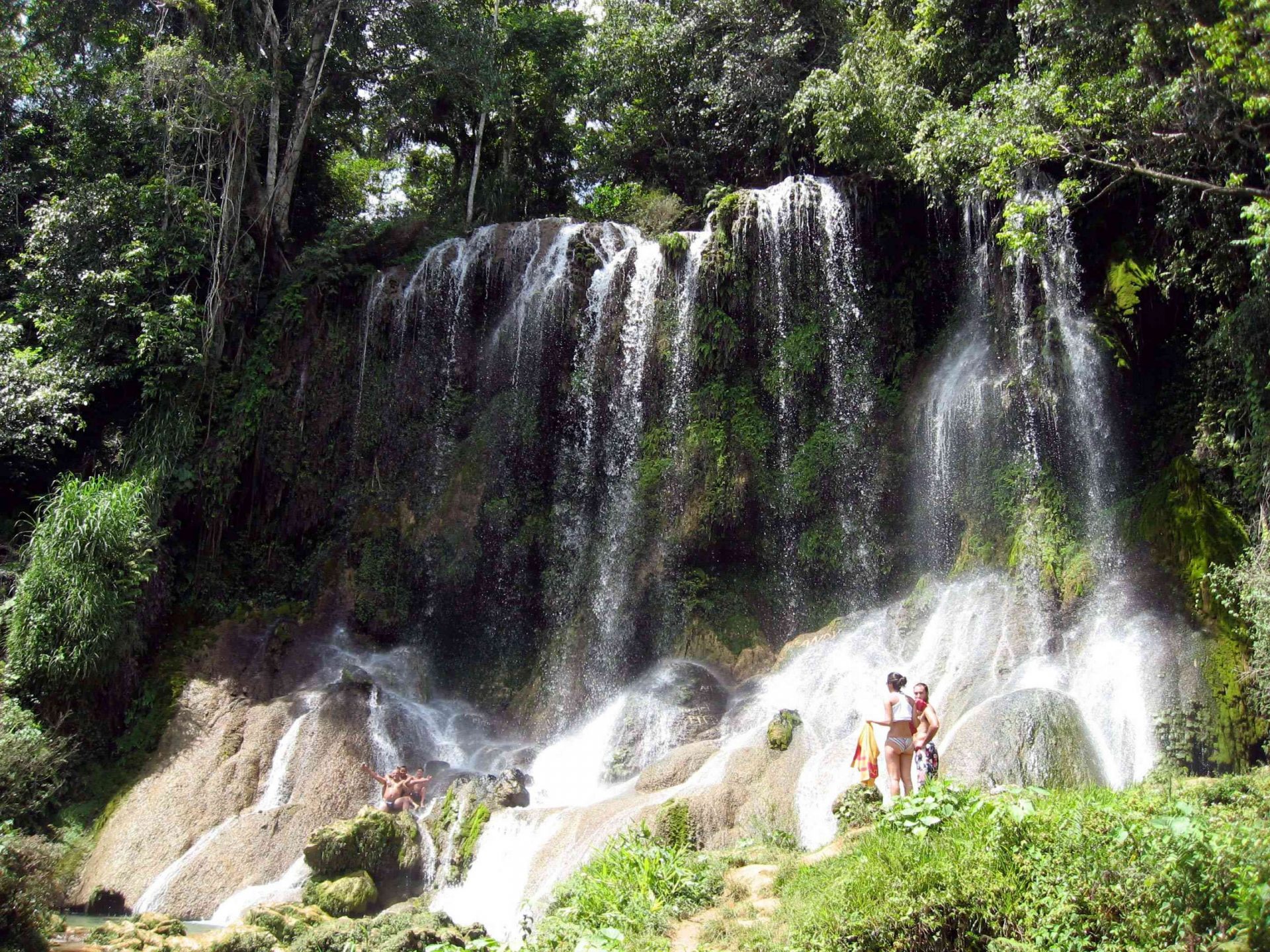 personas en la base de las cascadas que fluyen desde un bosque de altos árboles verdes y grandes rocas en la terminación de las cascadas en el Parque Nacional Desembarco del Granma
