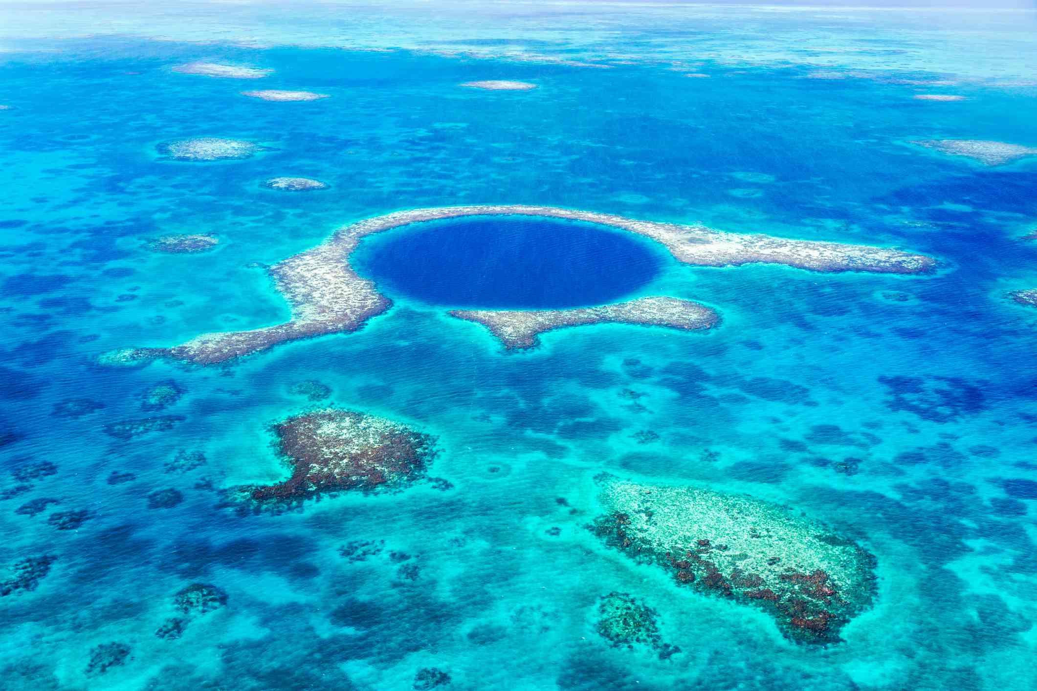 Un círculo de agua azul más oscuro en el océano revela una cueva submarina, rodeada de arrecifes blancos