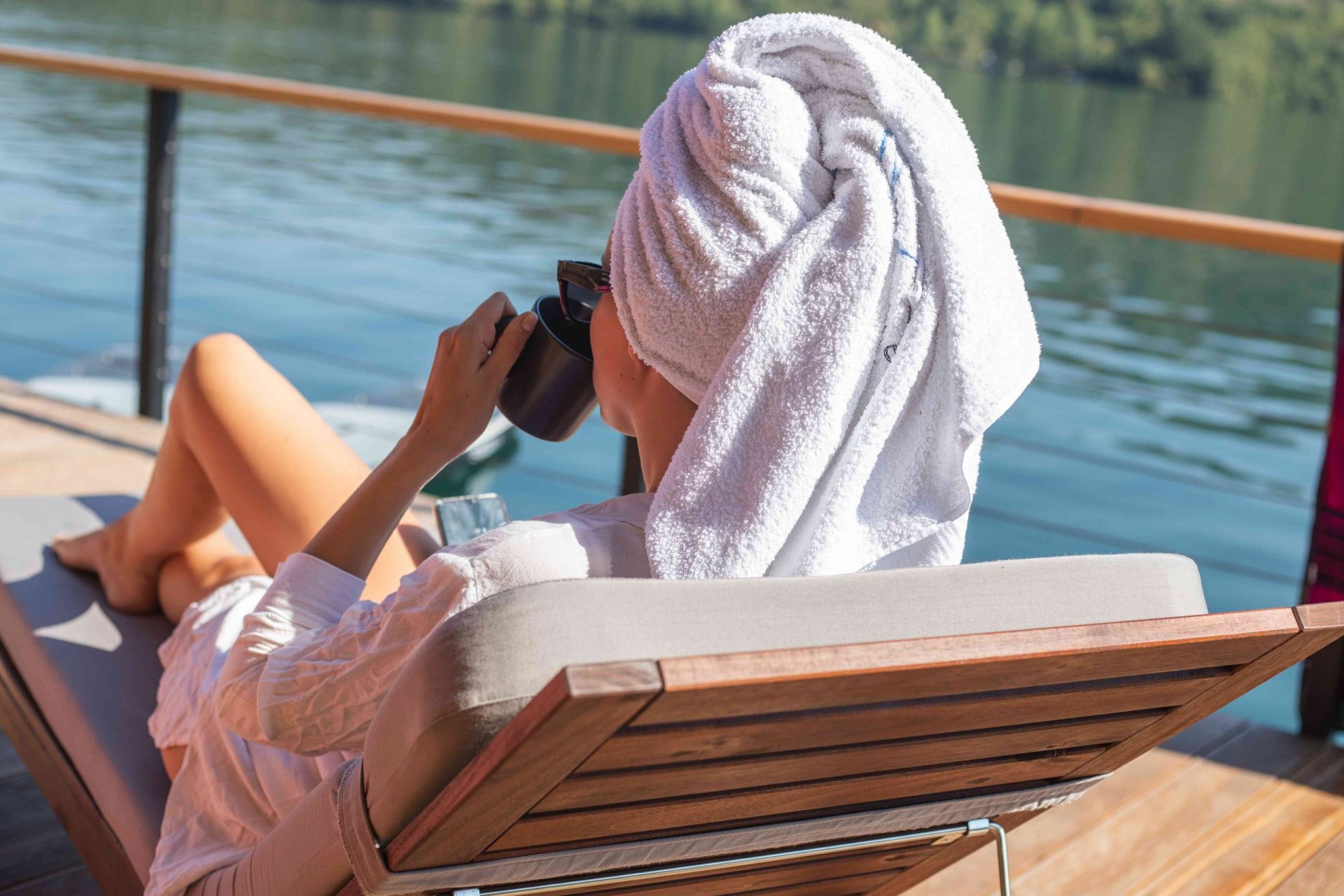 una mujer se relaja en una tumbona en un lago mientras se envuelve el pelo en una toalla para acondicionarlo en profundidad