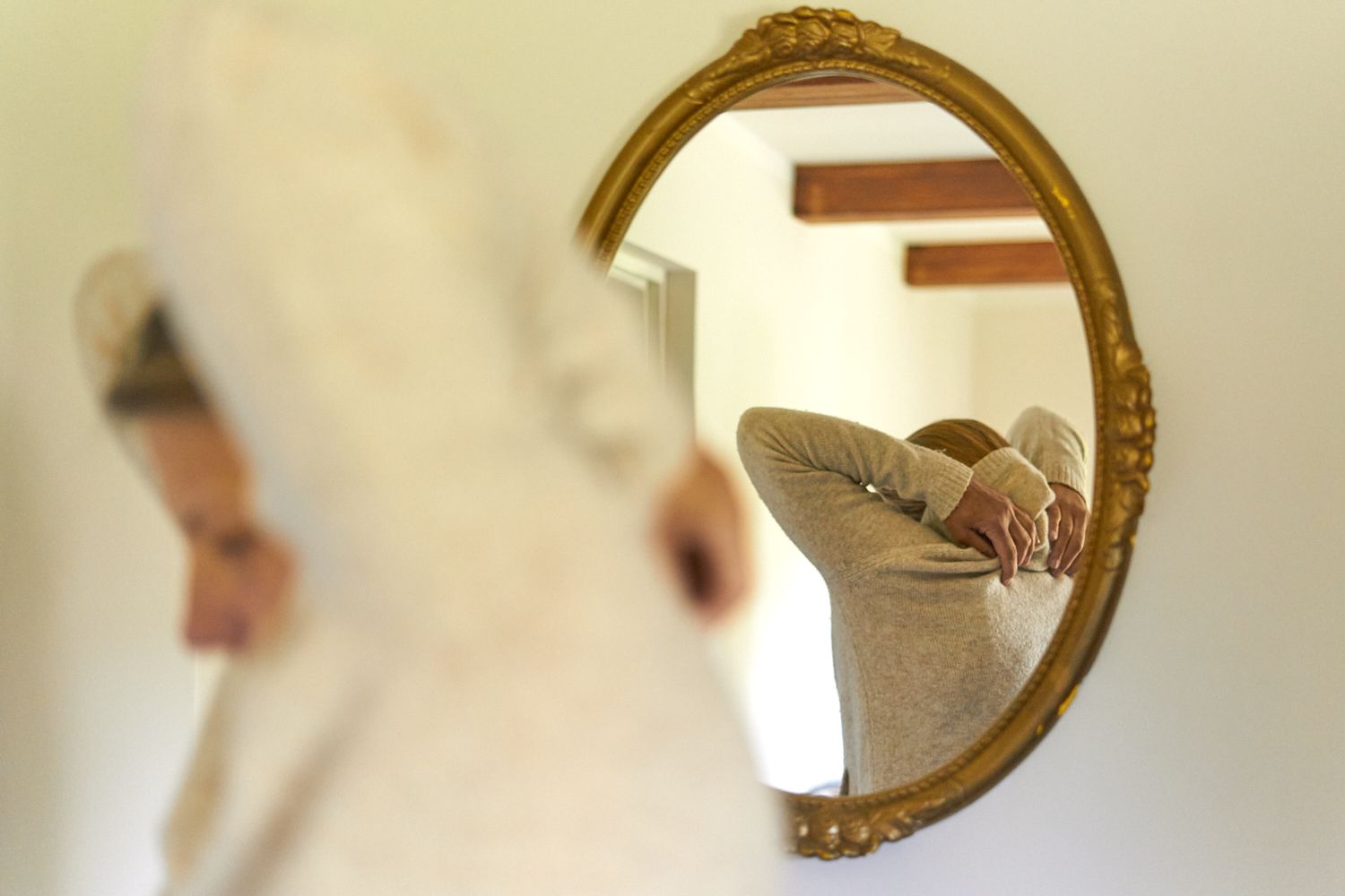 reflejo de una persona en un espejo ovalado dorado quitándose un jersey de color crema
