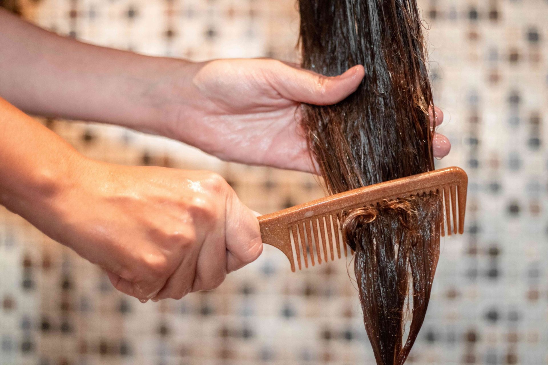 dos manos usan el peine marrón para pasar la mascarilla capilar por el pelo en la ducha alicatada