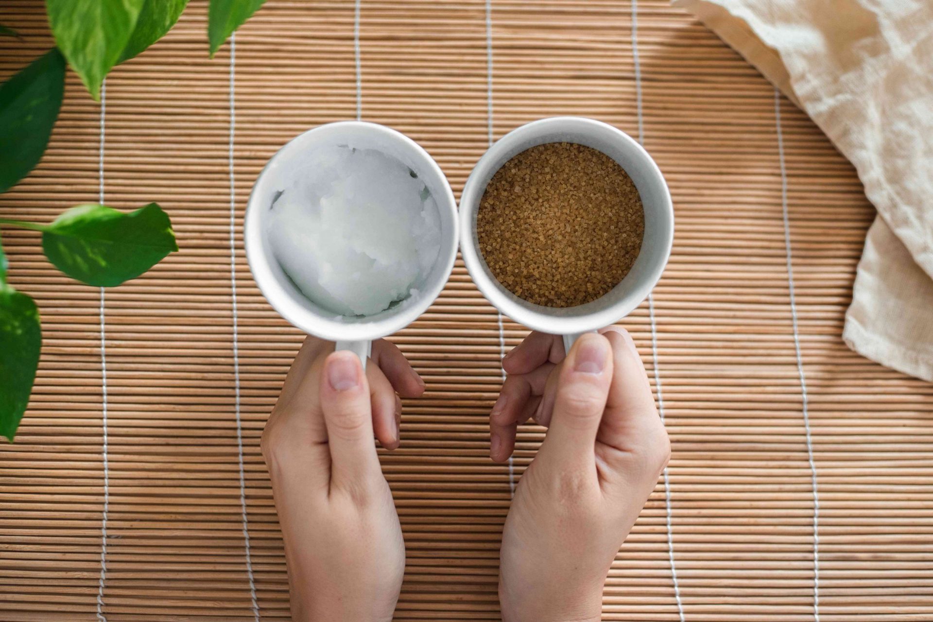 dos manos sostienen tazas de café llenas de aceite de coco sólido y azúcar moreno para crear el exfoliante