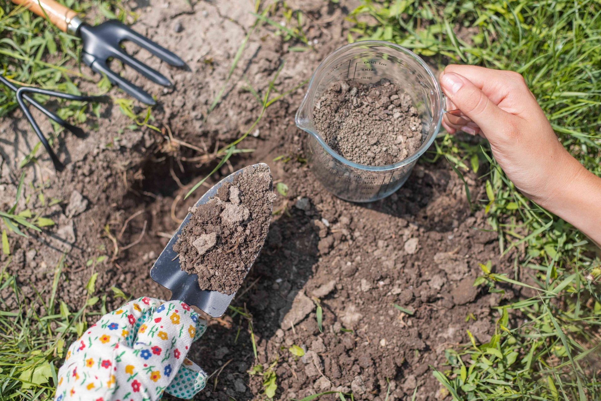 mano con herramientas de jardinería y vaso medidor recogiendo tierra para analizarla