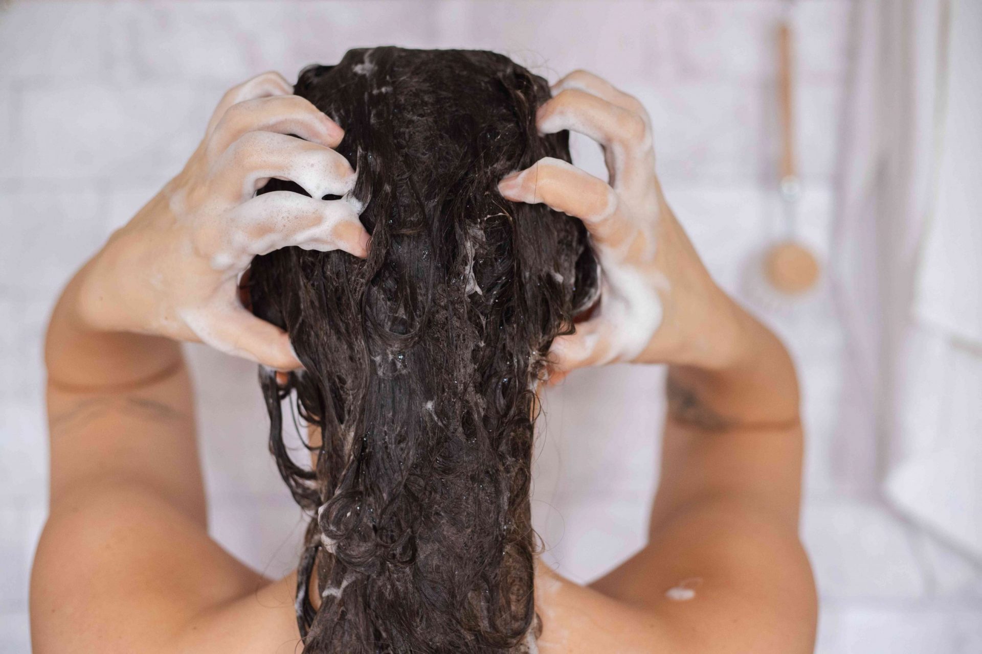 una mujer se frota el cuero cabelludo y el pelo con un champú espumoso en una ducha de azulejos blancos