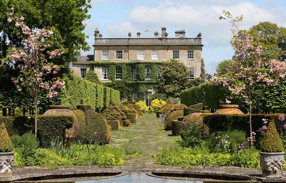 Vista de los clásicos jardines ingleses de High Grove del príncipe Carlos: dos árboles de flores rosas flanquean la entrada, seguidos de una variedad de setos verdes pulcramente esculpidos que conducen a la propiedad