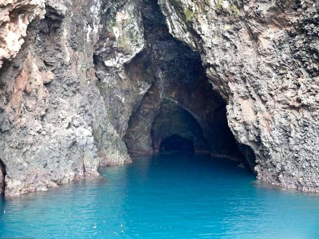 Una entrada a la cueva con paredes de roca irregular y un suelo cubierto de agua azul brillante