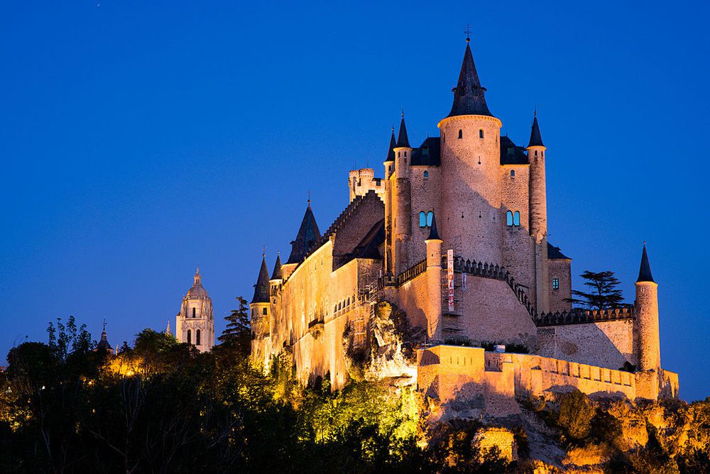 Vista del castillo del Alcázar de Segovia iluminado en su parte frontal e izquierda en el crepúsculo bajo un cielo azul claro