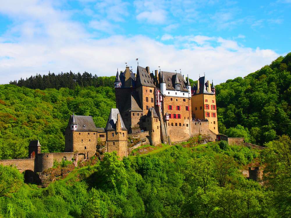 Burg Eltz, un castillo en una colina rodeado de un exuberante follaje verde bajo un cielo azul con nubes blancas