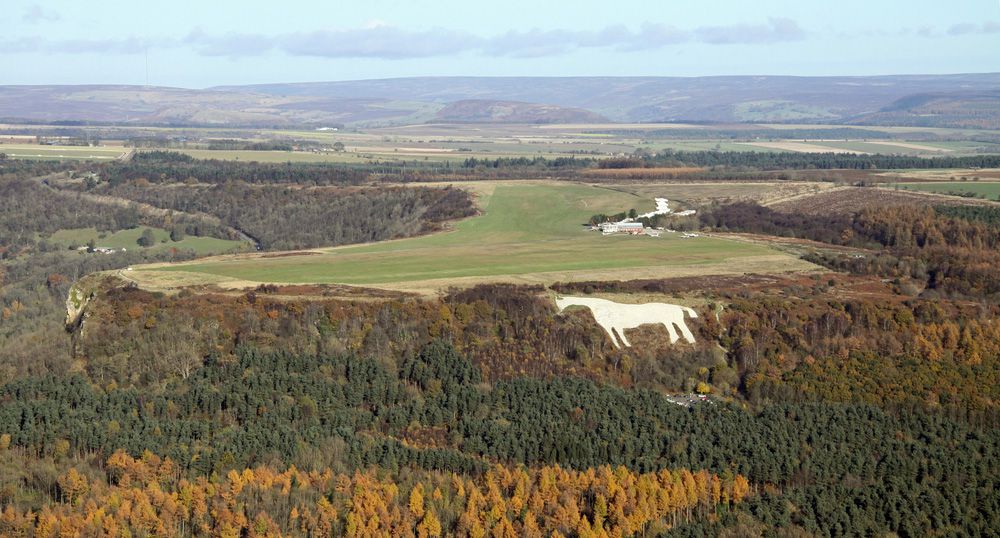 Vista aérea de la talla de piedra caliza de Kilburn White Horse en una colina verde rodeada de árboles dorados y verdes en las colinas circundantes bajo un cielo azul brillante