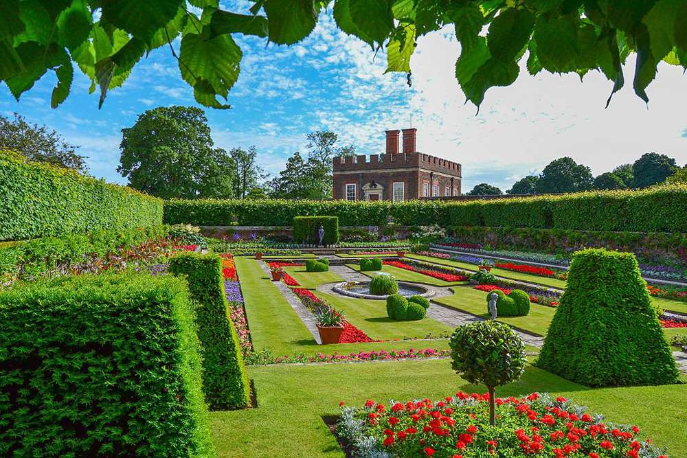 Jardines ingleses clásicos bajo un cielo azul cubiertos de césped verde pulcramente recortado, setos verdes esculpidos y parterres de flores rojas frente al Palacio de Hampton Court