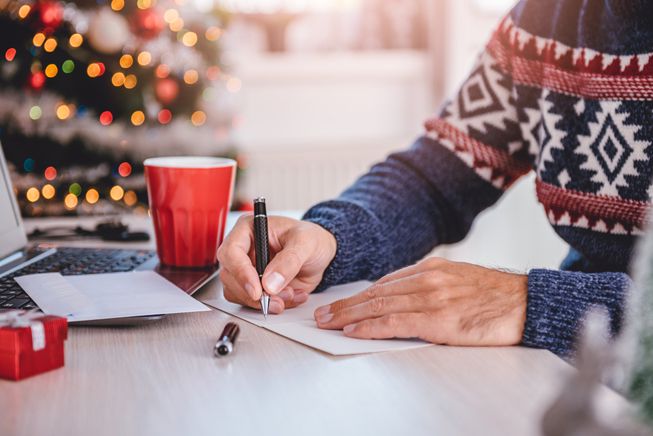 Una persona con jersey escribe en una tarjeta de Navidad en un escritorio cerca de un árbol de Navidad