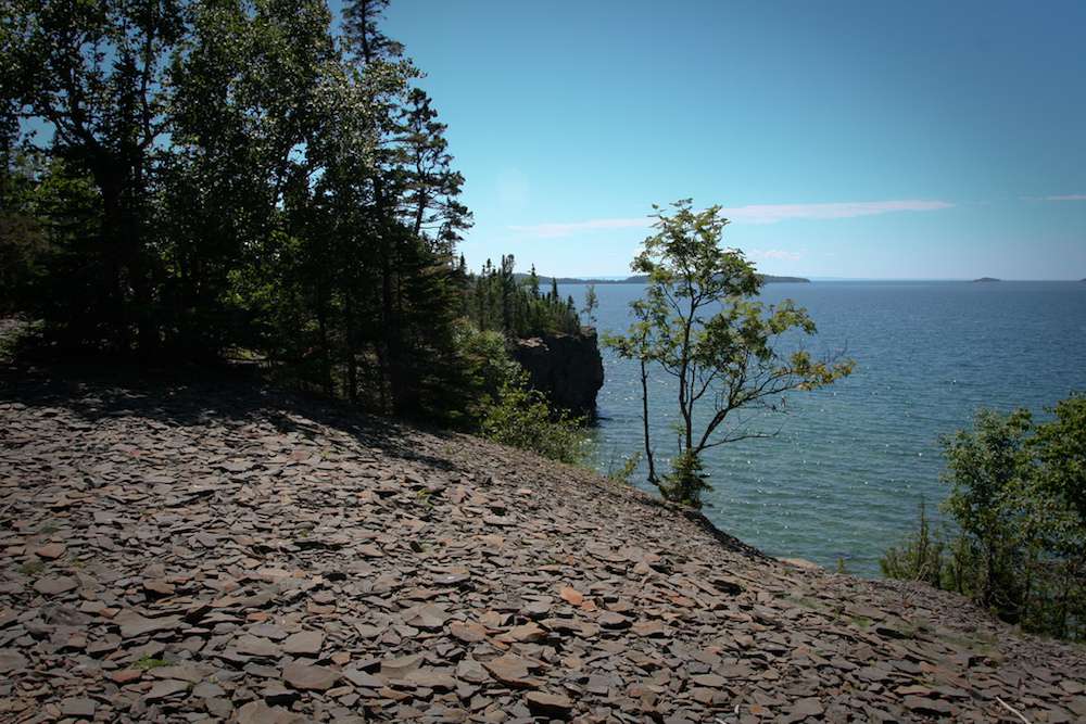 Islote de Plata en el Parque Provincial del Gigante Durmiente, con árboles y rocas en la orilla y el Lago Superior de fondo bajo un cielo azul
