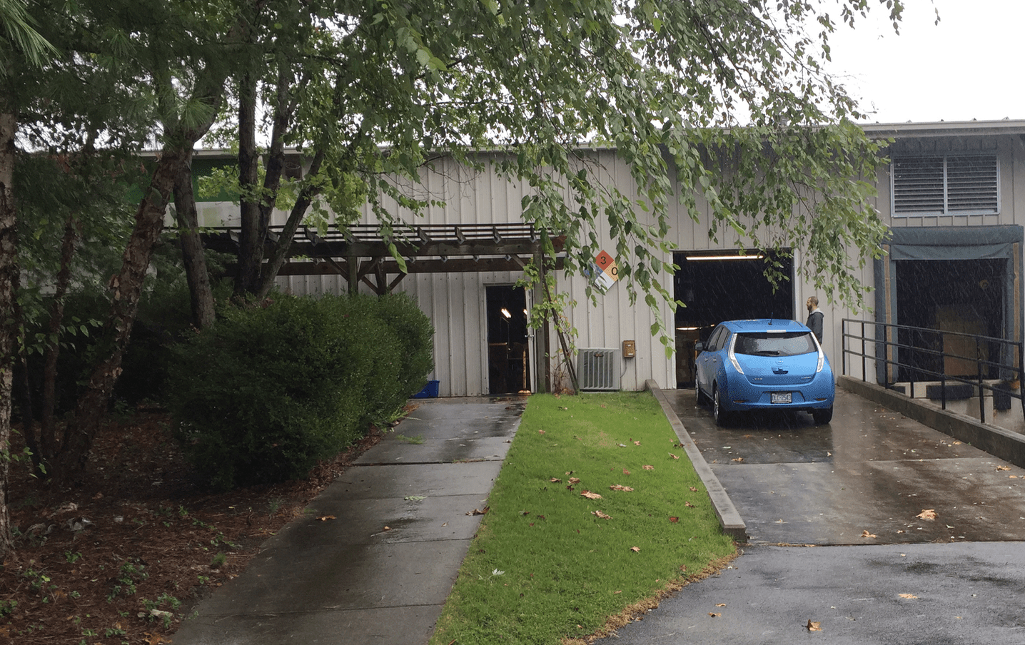 Un edificio de tejado plano con un coche azul entrando y un árbol a la izquierda