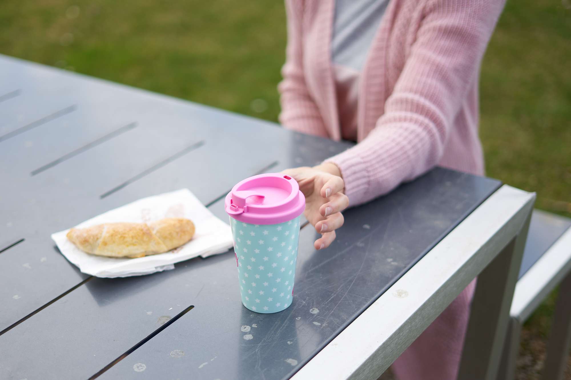 una mujer con rebeca rosa en una mesa de picnic coge una taza de café reutilizable rosa y verde azulado
