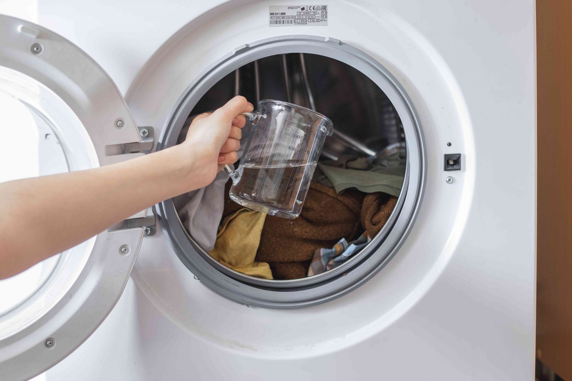 abre la puerta de la lavadora con la mano sujetando una jarra de vinagre para verterla sobre la ropa sucia