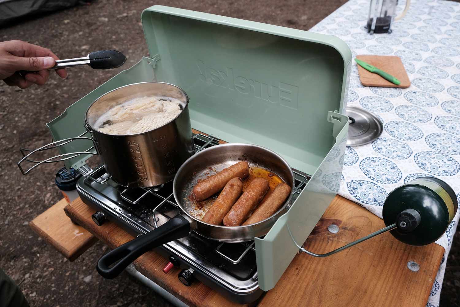 un hombre cocina salchichas en un hornillo de camping en una mesa de picnic
